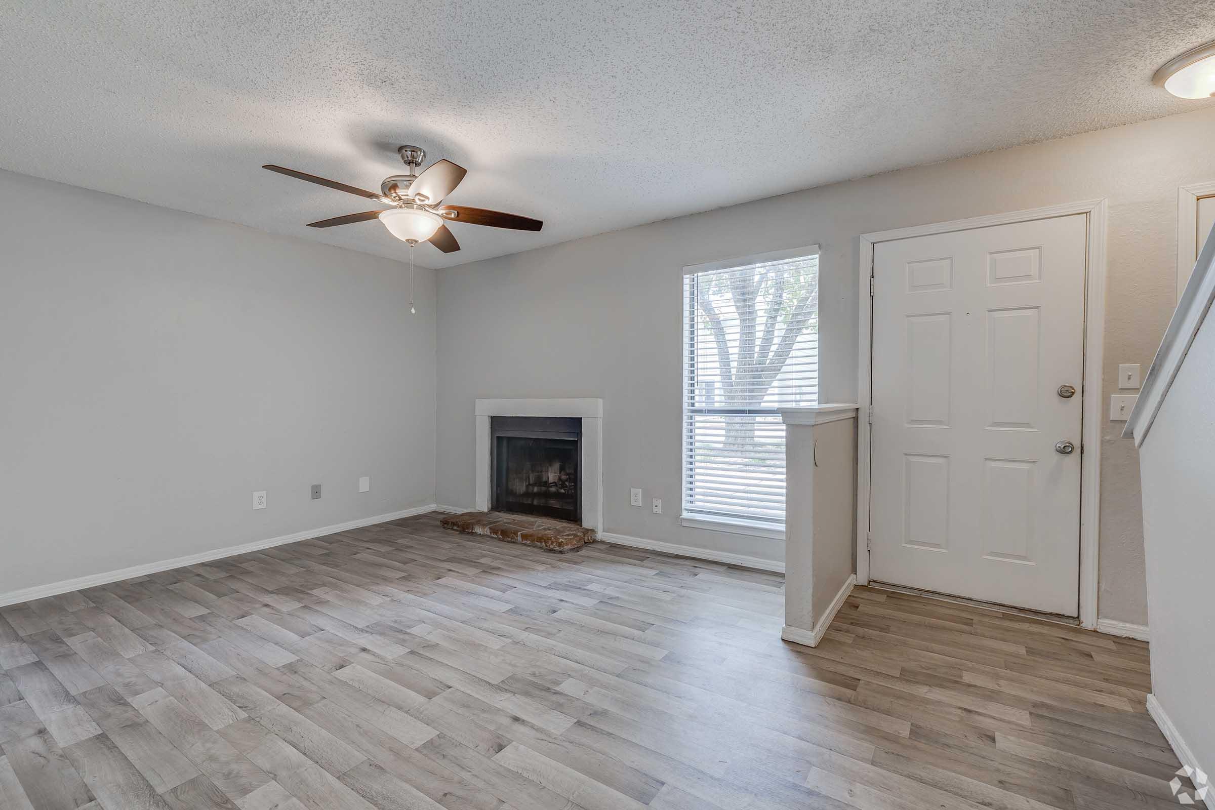 A modern, empty living room featuring a ceiling fan, wooden flooring, and a fireplace. Natural light comes from a large window with blinds, alongside a front door with a small entryway. The walls are painted in a light color, creating a clean and airy atmosphere.