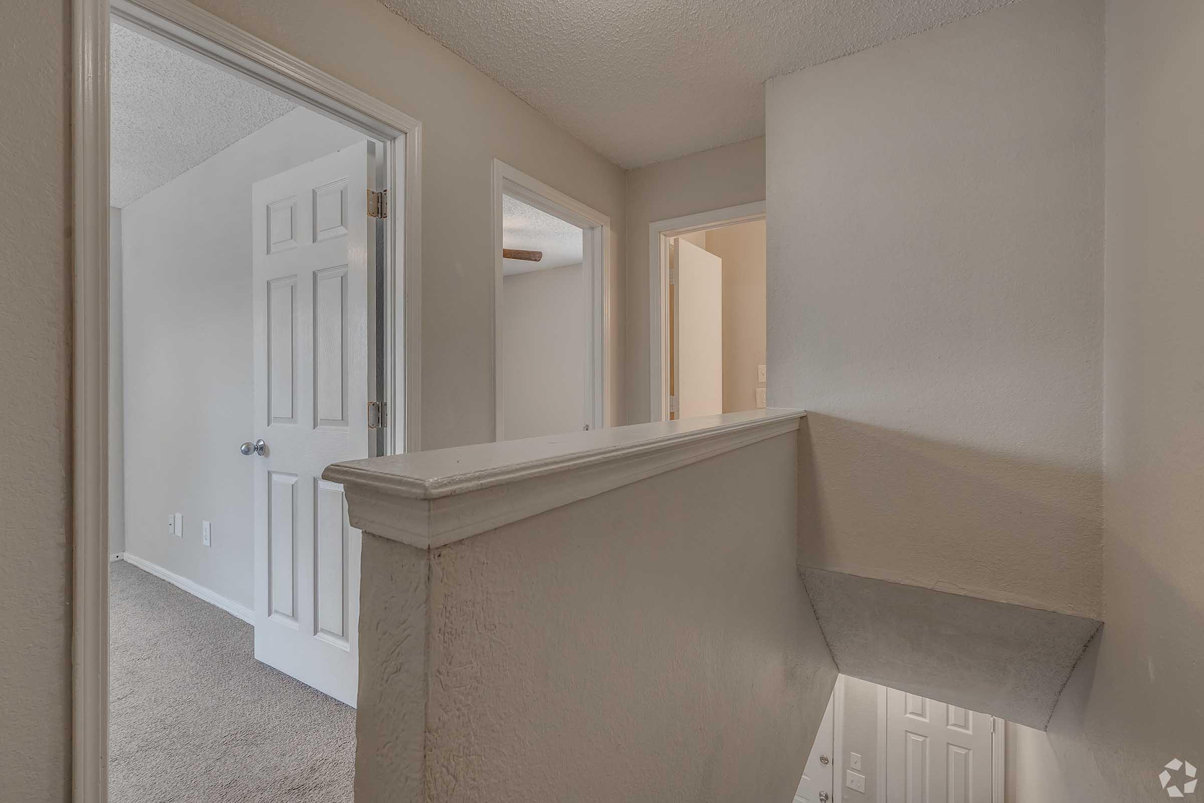 Interior view of a hallway with freshly painted walls and light-colored carpet. Visible doors lead to adjacent rooms, and a staircase descends at the end of the hallway. The lighting is soft, enhancing the warm atmosphere of the space.