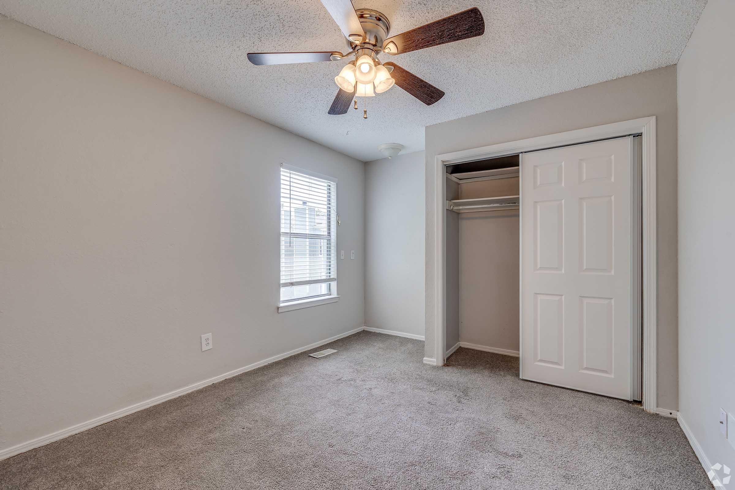 A light gray carpeted bedroom featuring a ceiling fan with wooden blades, a window with white blinds, and a closet with bi-fold doors. The walls are painted a neutral gray, and the room has natural light coming in from the window.