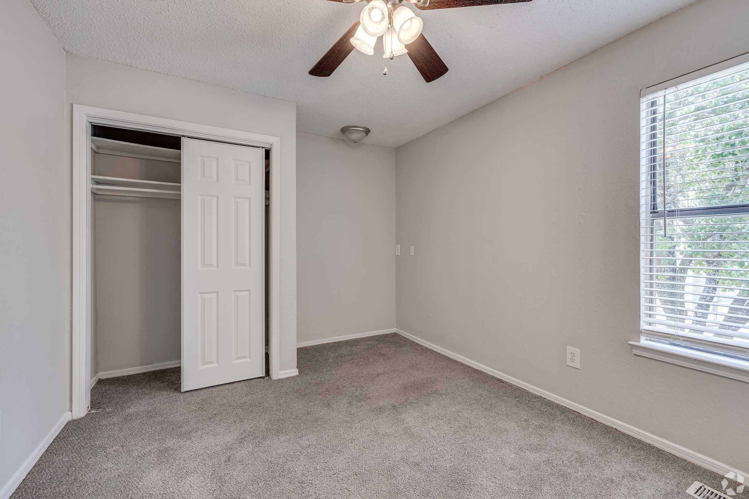 A small, empty bedroom with light gray walls and carpet. A ceiling fan is visible, and there's a window with blinds letting in natural light. A closet with sliding doors is on one side, while the opposite wall features a single electrical outlet. The overall atmosphere is clean and inviting.