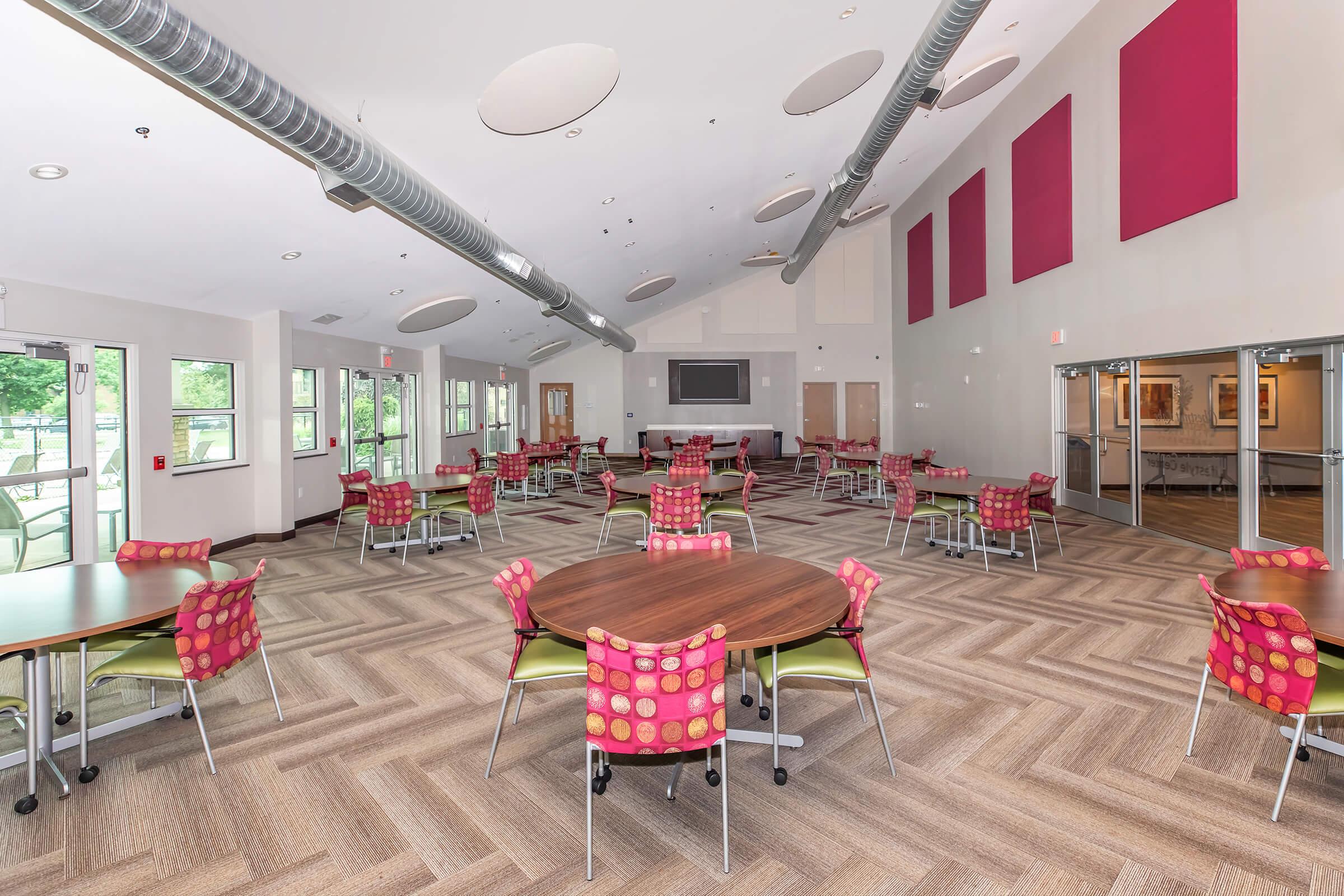 A spacious community room featuring multiple round tables with colorful chairs, large windows allowing natural light, a television screen mounted on the wall, and a modern, welcoming decor. The flooring has a herringbone pattern, enhancing the room's contemporary feel.