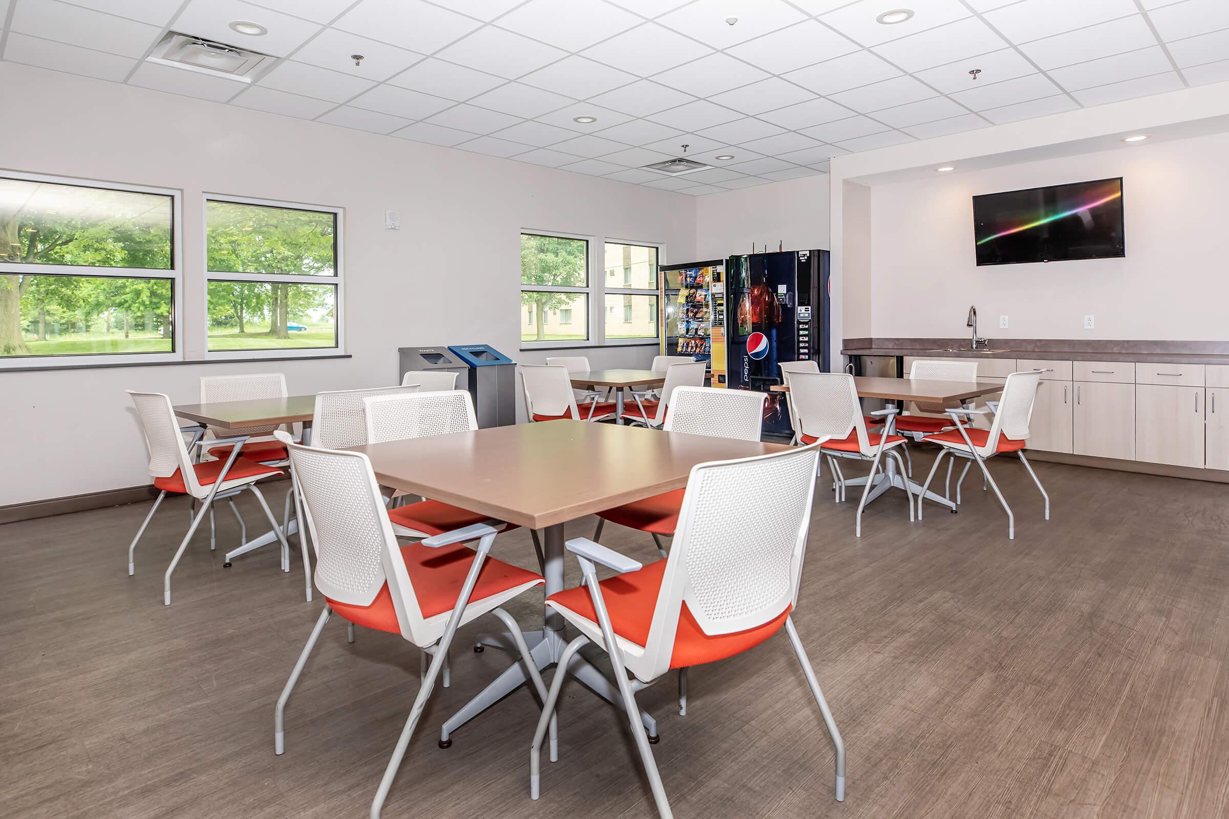 A modern communal room featuring several light-colored tables surrounded by white and orange chairs. There's a vending machine in the corner, and large windows provide a view of green outdoor space. A television hangs on the wall, and the overall design is bright and inviting.