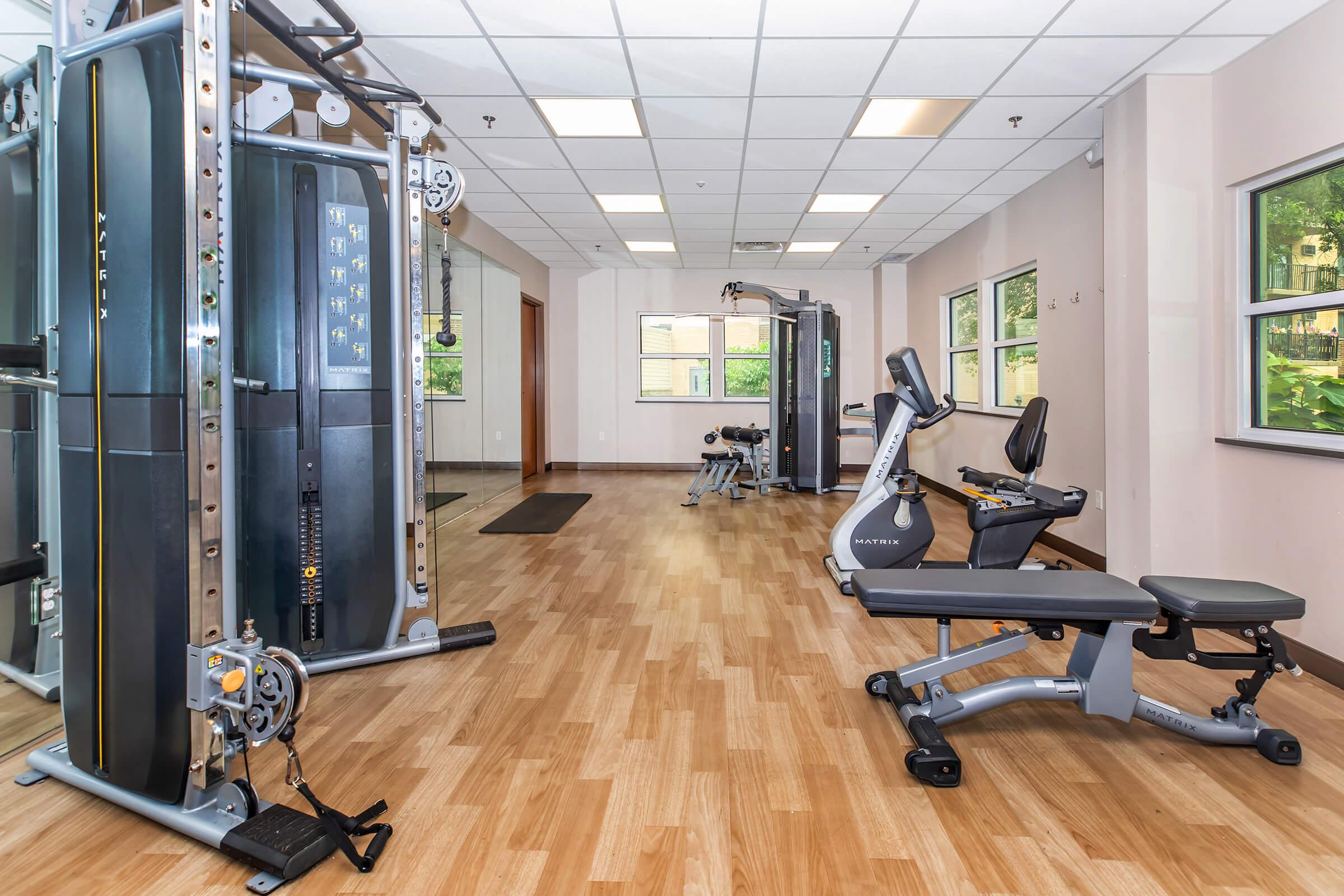 A modern gym interior featuring various fitness equipment. On the left, a multi-station weight machine, and on the right, a recumbent exercise bike. The space is well-lit with large windows, hardwood flooring, and a clean, organized layout, creating an inviting atmosphere for workouts.