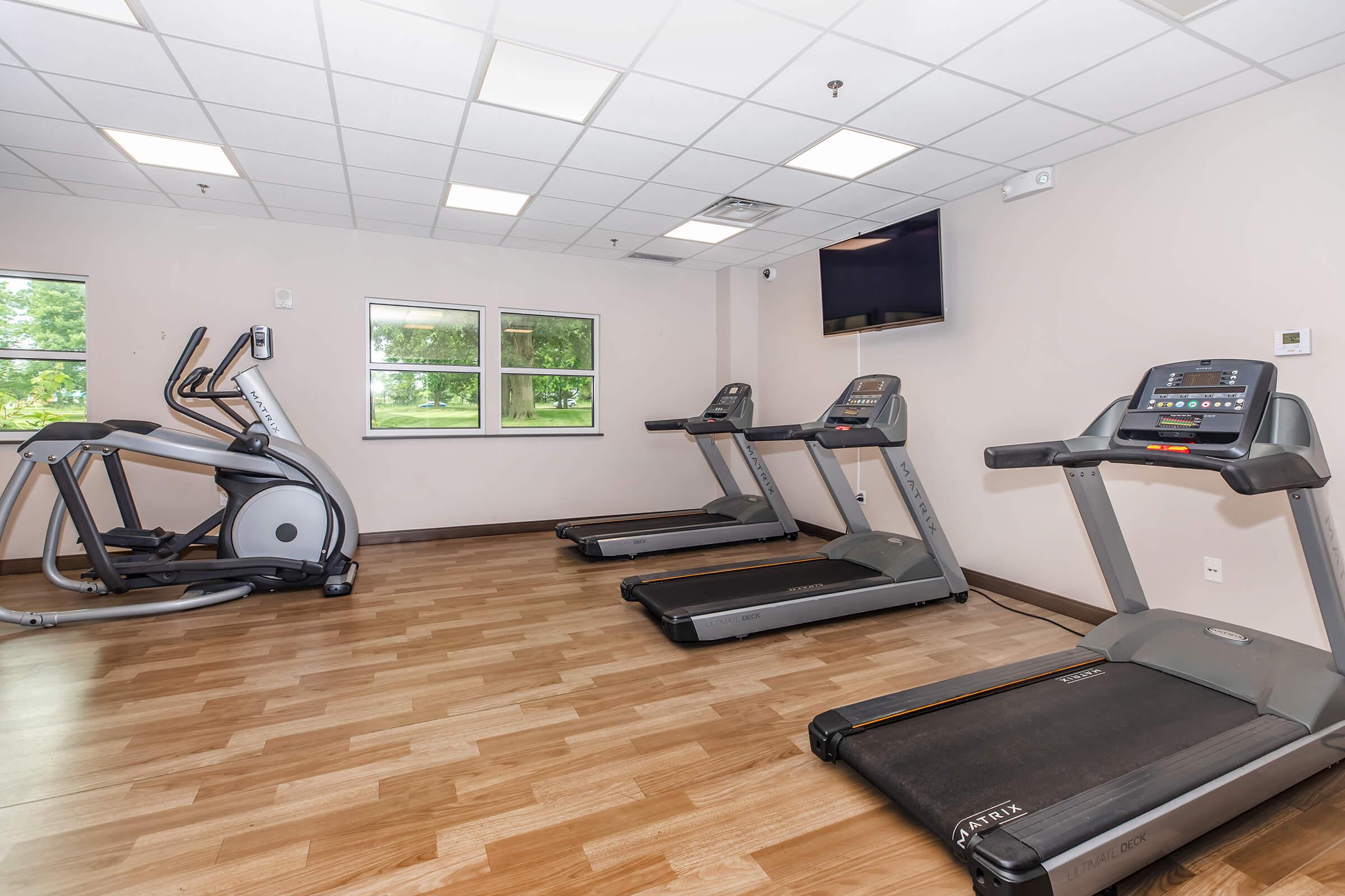 A well-lit indoor gym featuring two treadmills and an elliptical machine. The space has large windows allowing natural light, a flat-screen TV mounted on the wall, and a wooden floor.