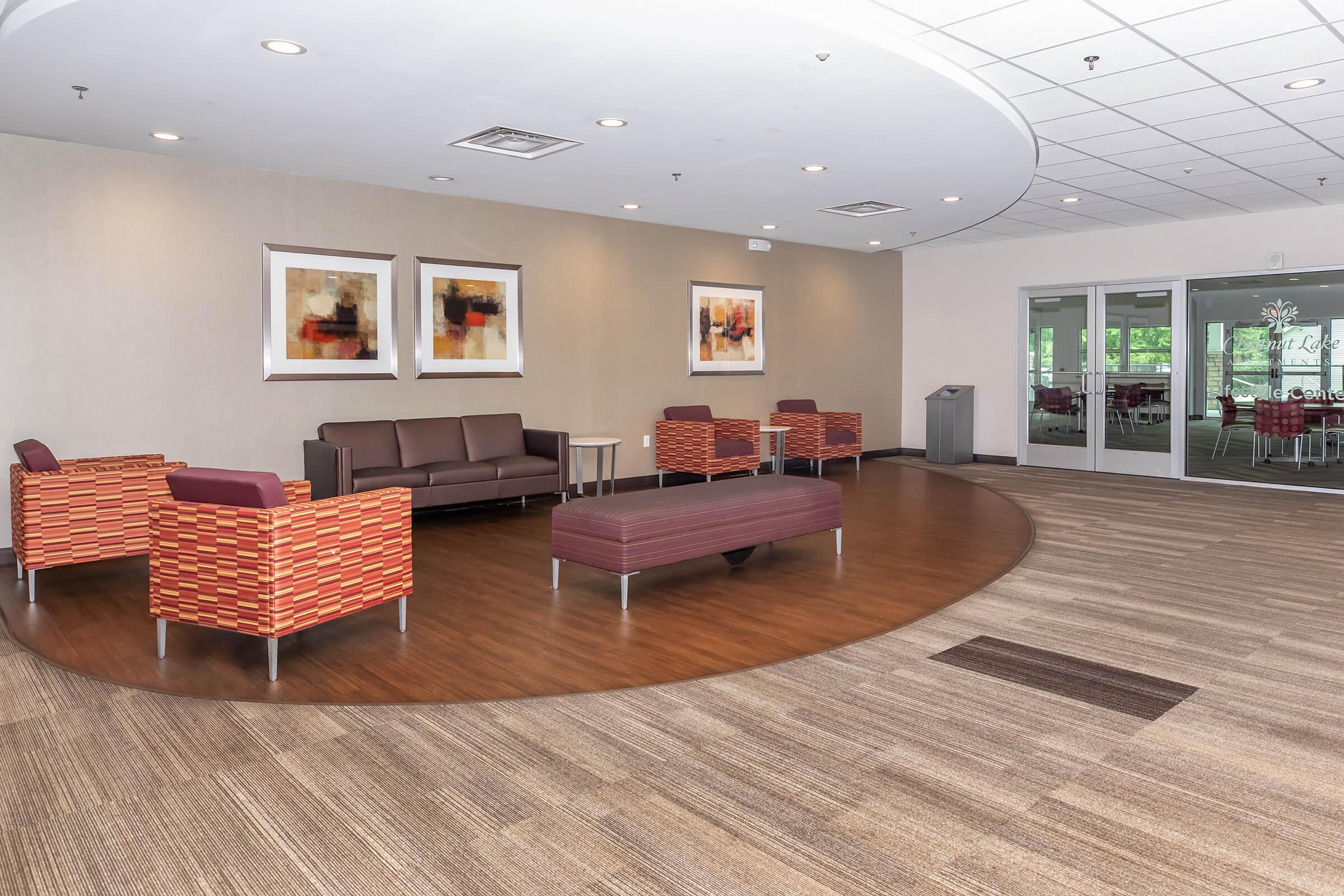 Interior of a modern lounge area with a curved layout. The space features a brown couch and patterned chairs arranged around a central purple bench. Light-colored walls and a neutral-toned floor create a warm atmosphere. Abstract art is displayed on the walls, and large glass doors lead to an outdoor area.