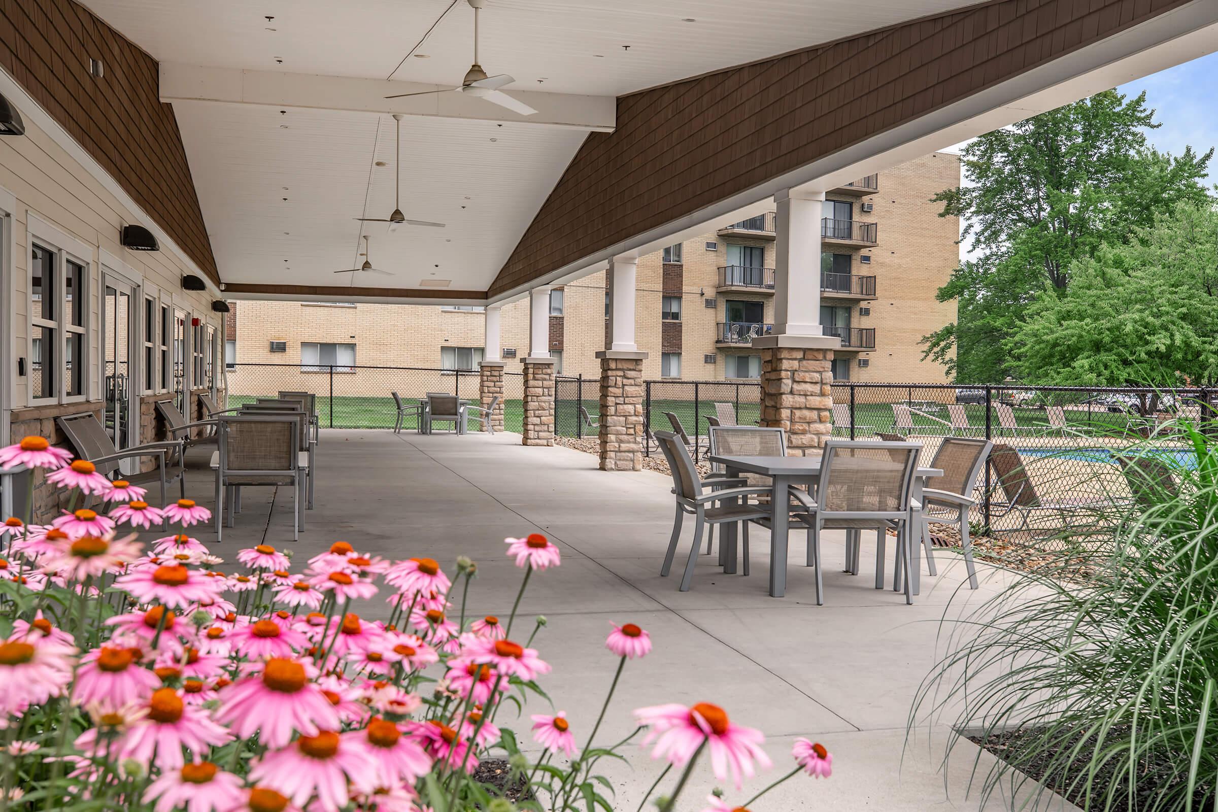 A covered patio area featuring several tables and chairs, bordered by blooming pink flowers. In the background, there are residential buildings and greenery, creating a serene outdoor setting perfect for relaxation. Ceiling fans are installed for comfort.