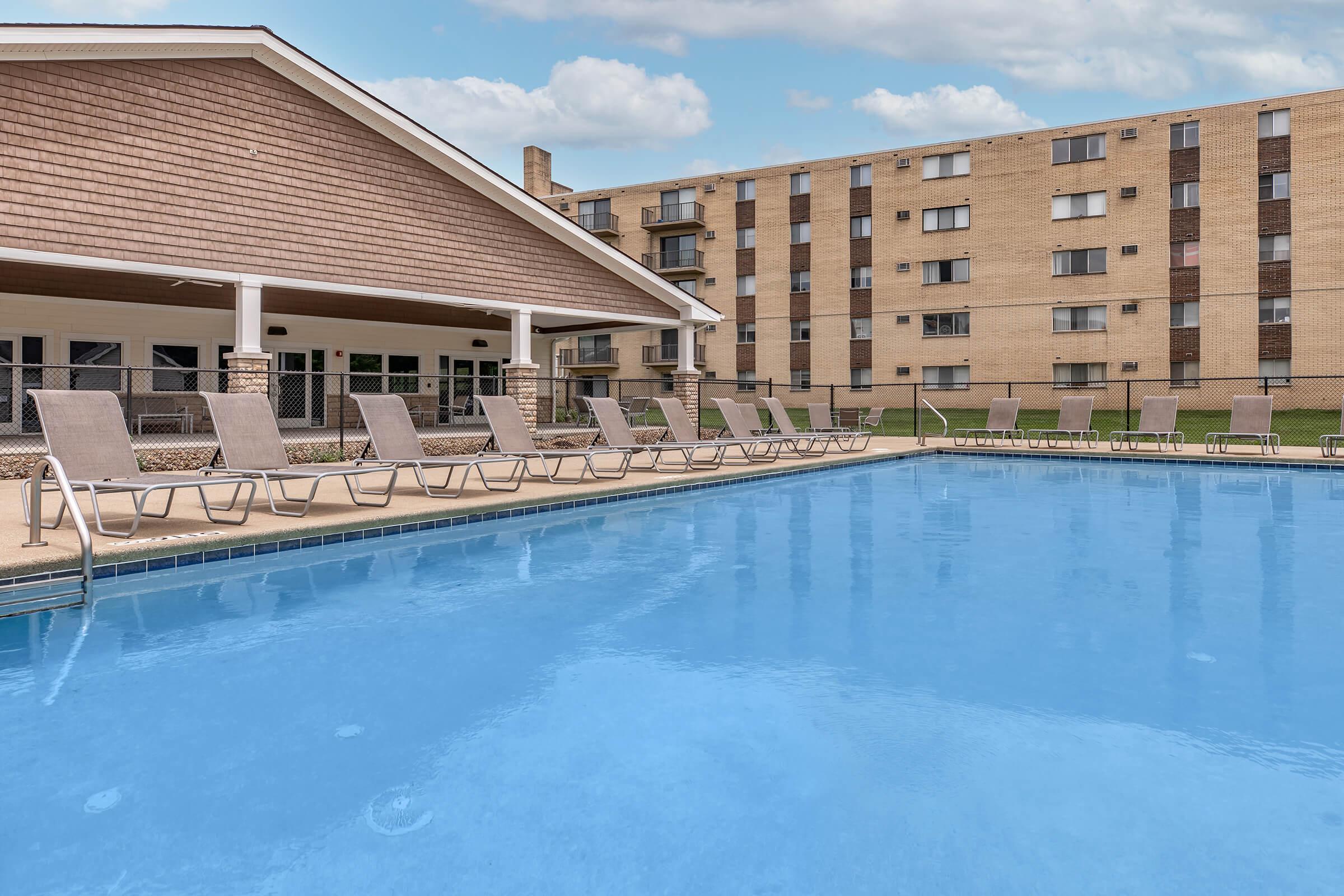 A clear blue swimming pool with several lounge chairs lined up along the edge. In the background, there is a large building with multiple floors and windows. The scene is set under a partly cloudy sky, creating a relaxing outdoor atmosphere.