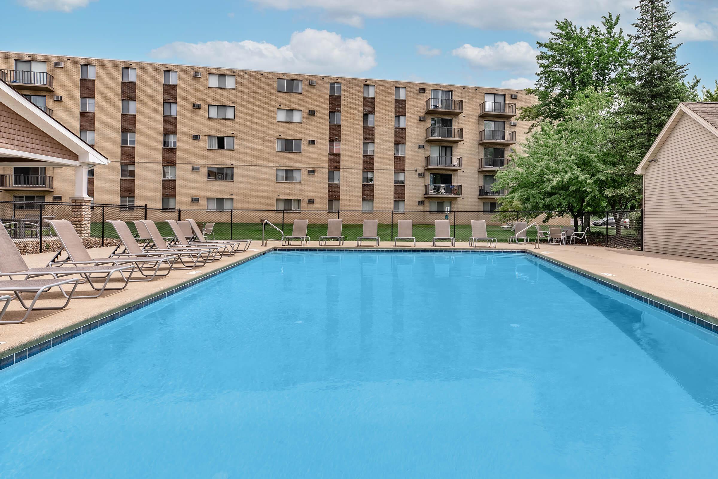 A clear blue swimming pool surrounded by lounge chairs, set against a backdrop of a multi-story apartment building and greenery. The sky is partially cloudy, creating a serene atmosphere for relaxation.