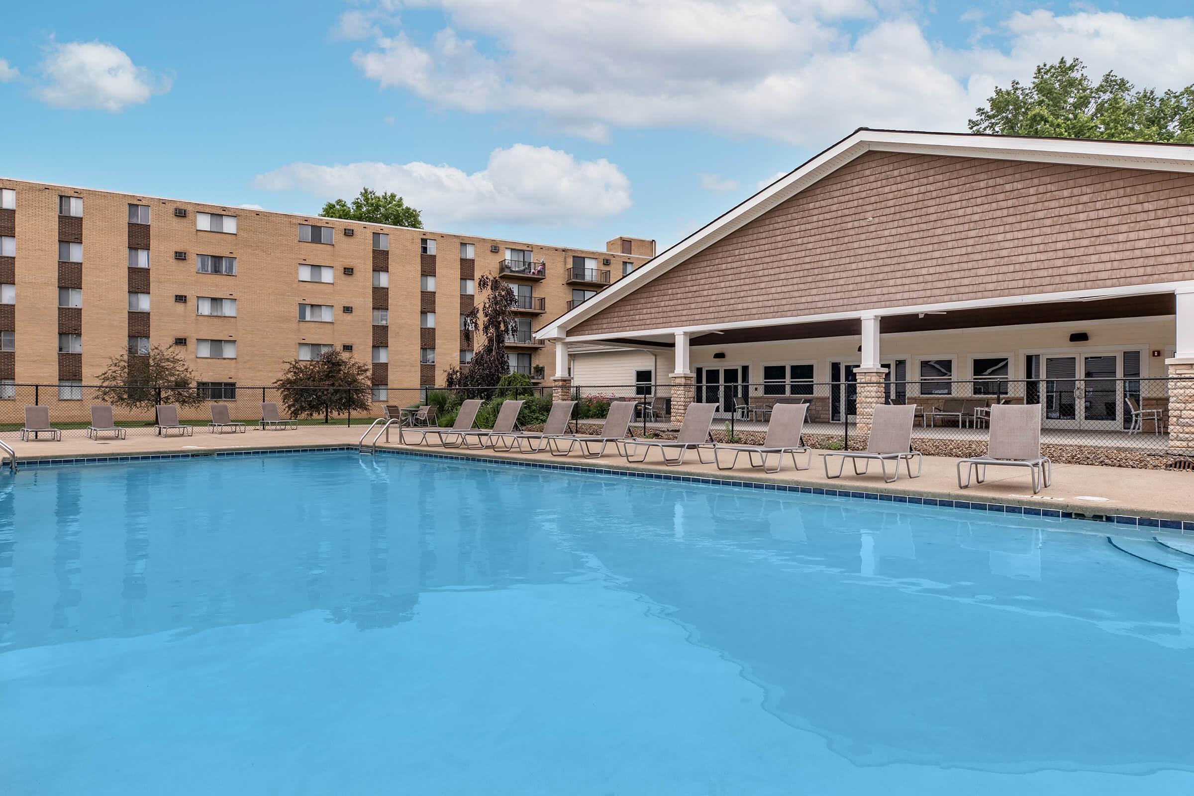 A clear blue swimming pool with lounge chairs positioned around it. In the background, there is a beige apartment building with multiple floors and windows. The sky is partly cloudy, contributing to a bright, inviting atmosphere.