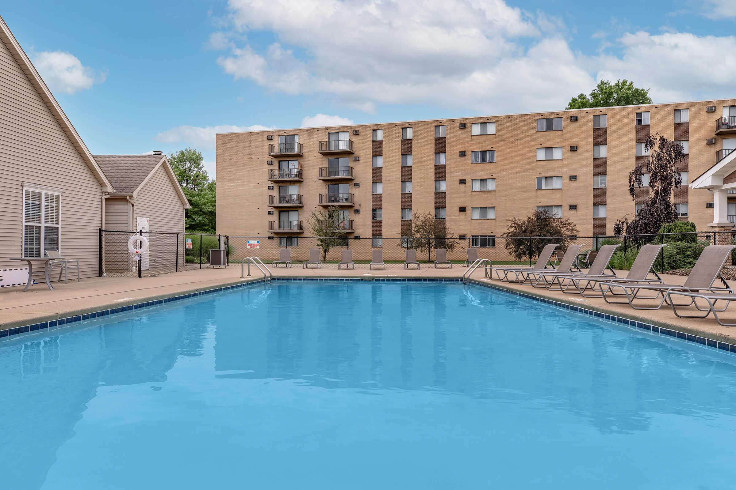 A clear blue swimming pool with lounge chairs around it, adjacent to a multi-story apartment building. The sky is partly cloudy, and there are trees in the background, creating a relaxed outdoor atmosphere.