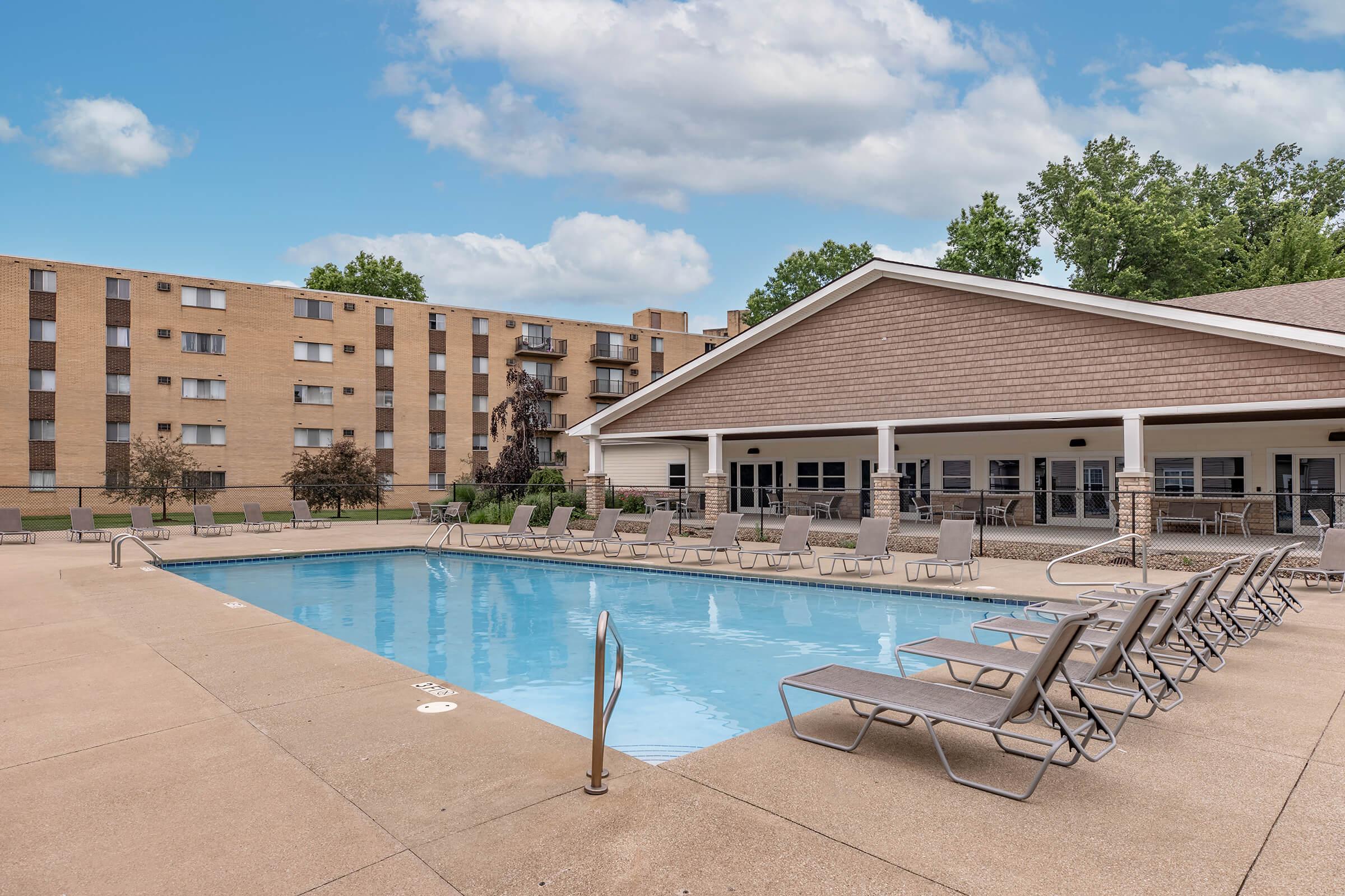A clean outdoor swimming pool surrounded by lounge chairs, with a modern clubhouse in the background. The area features landscaped greenery and a clear blue sky, creating a welcoming atmosphere for relaxation and leisure.