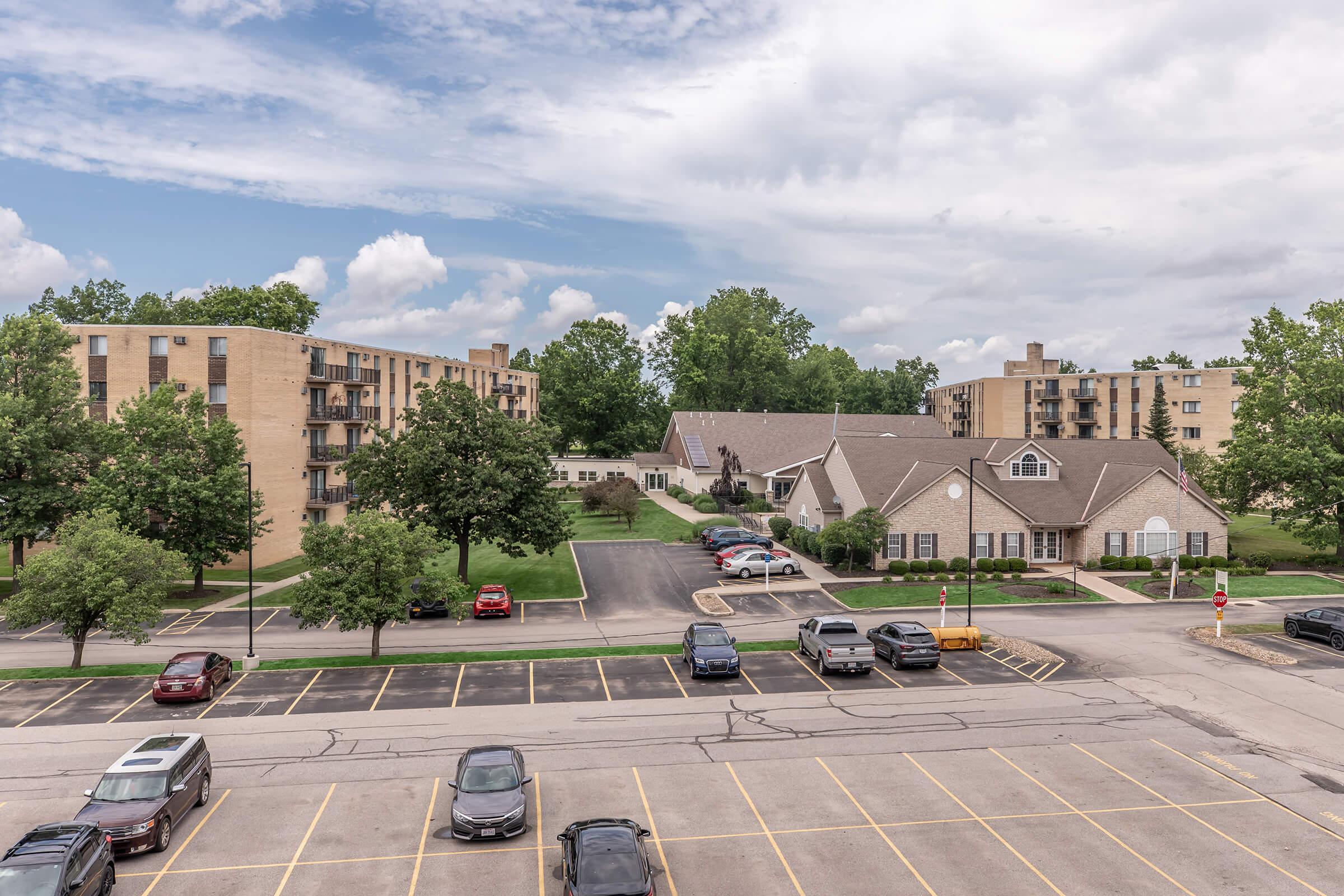 View of an apartment complex with several multi-story buildings surrounded by green trees and a cloudy sky. The foreground features a parking lot with multiple cars parked in designated spaces, while the background includes a residential office or community center.