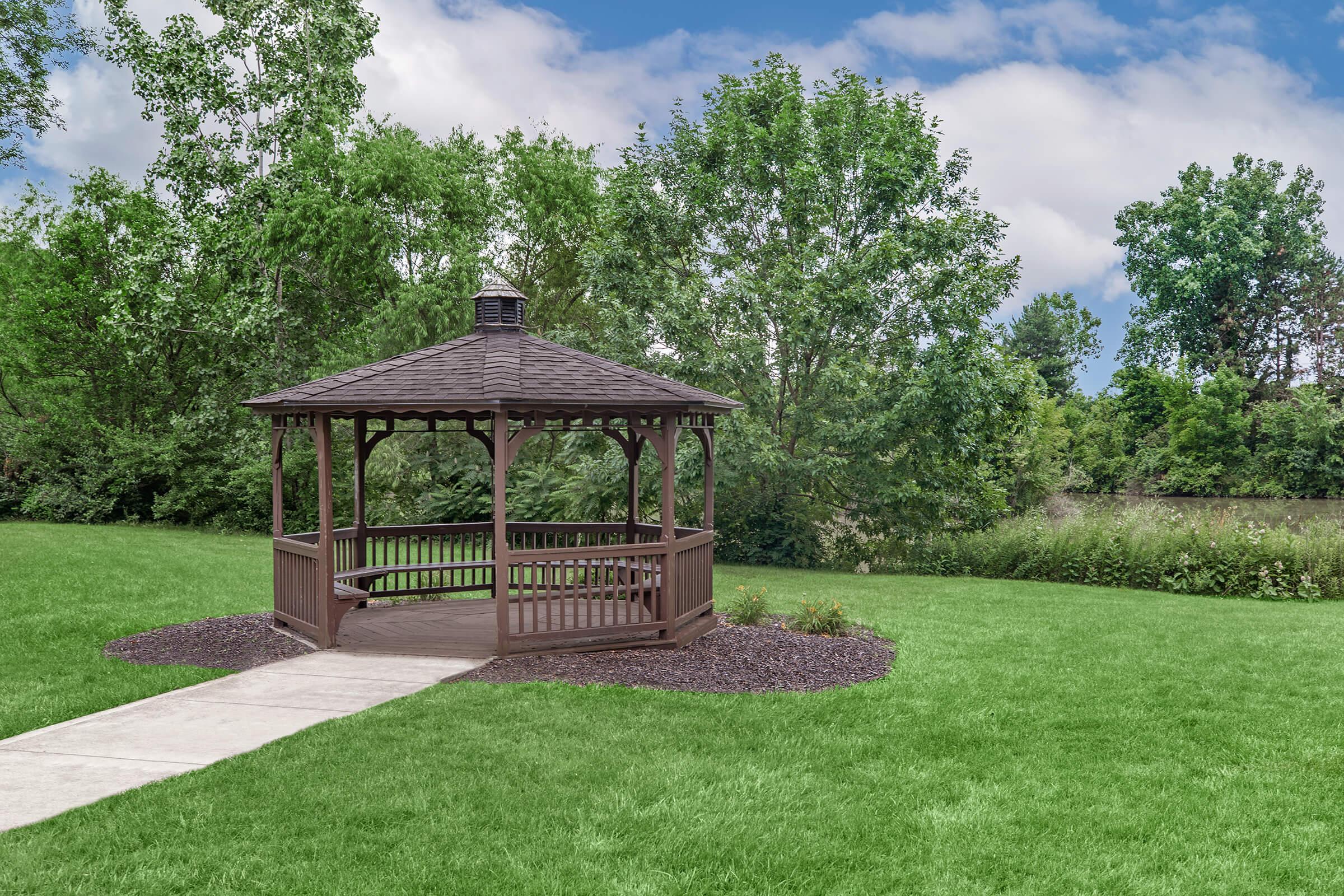 A wooden gazebo with a pointed roof, situated on a well-maintained grassy area. Surrounding the gazebo are trees and shrubs, under a partly cloudy blue sky. A concrete walkway leads up to the structure, creating an inviting spot for relaxation or gatherings.