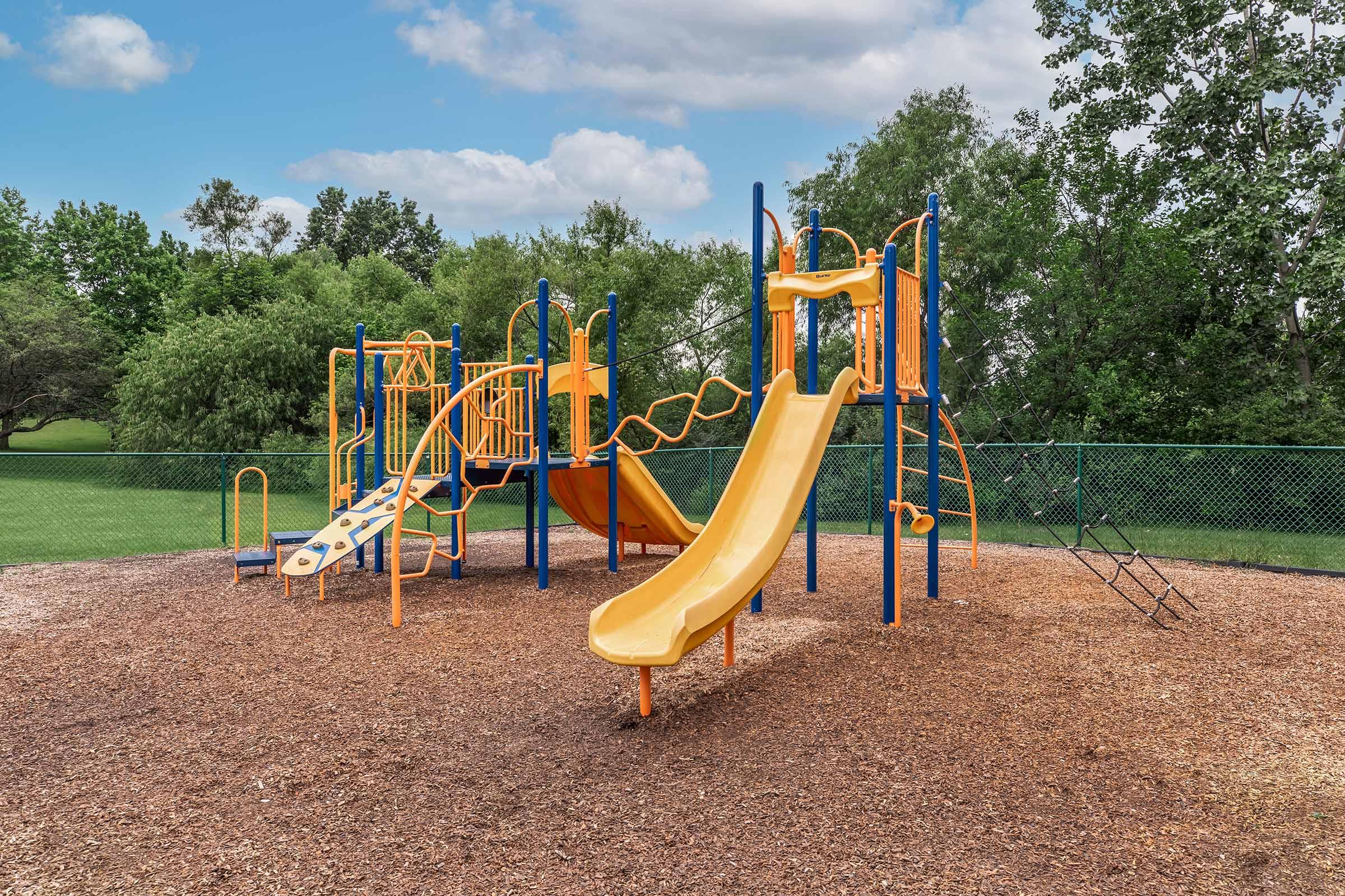 A colorful children's playground featuring two yellow slides, climbing structures, and safety railings, set on a bed of wood chips. In the background, there are green trees and a fence, creating a sunny and inviting outdoor play area.