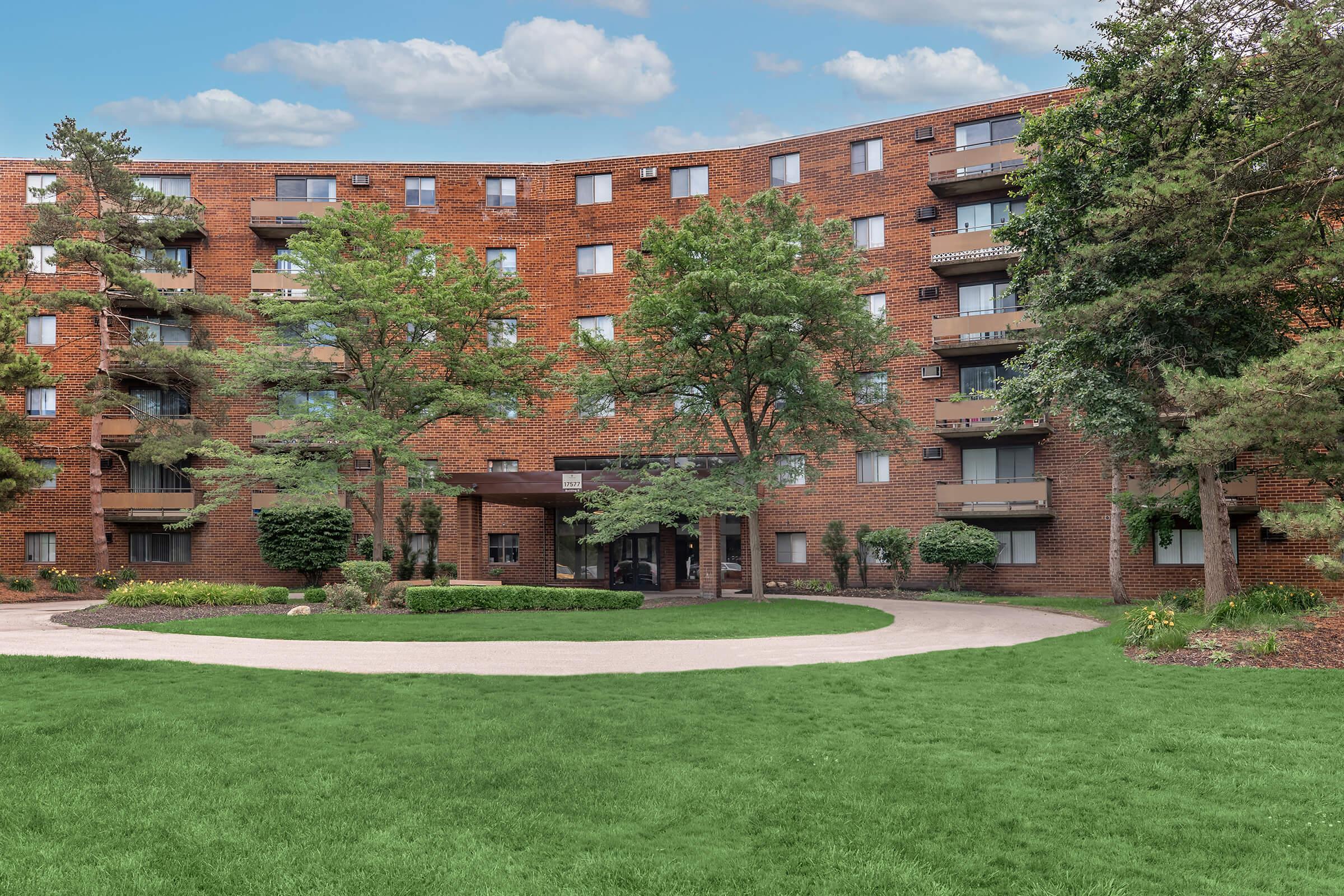 Landscape view of a brick apartment building surrounded by lush greenery. The structure features multiple balconies and large windows. A paved pathway curves through a neatly manicured lawn leading to the entrance. The scene is set against a clear blue sky with some clouds.
