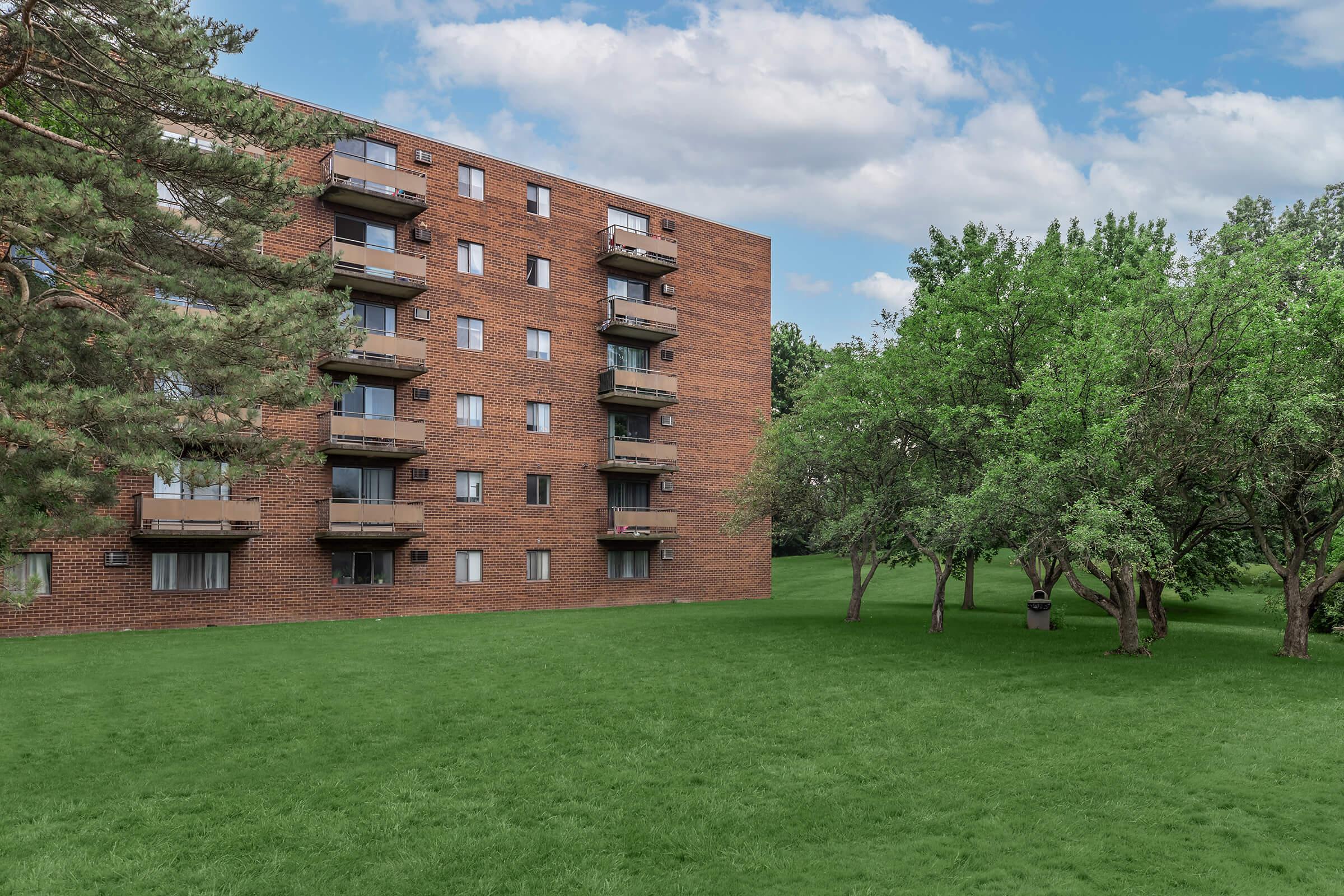 A red brick apartment building with multiple balconies overlooks a lush green lawn. Nearby, several trees provide shade, and a trash can is visible in the background. The sky is partly cloudy, adding to the serene outdoor setting.