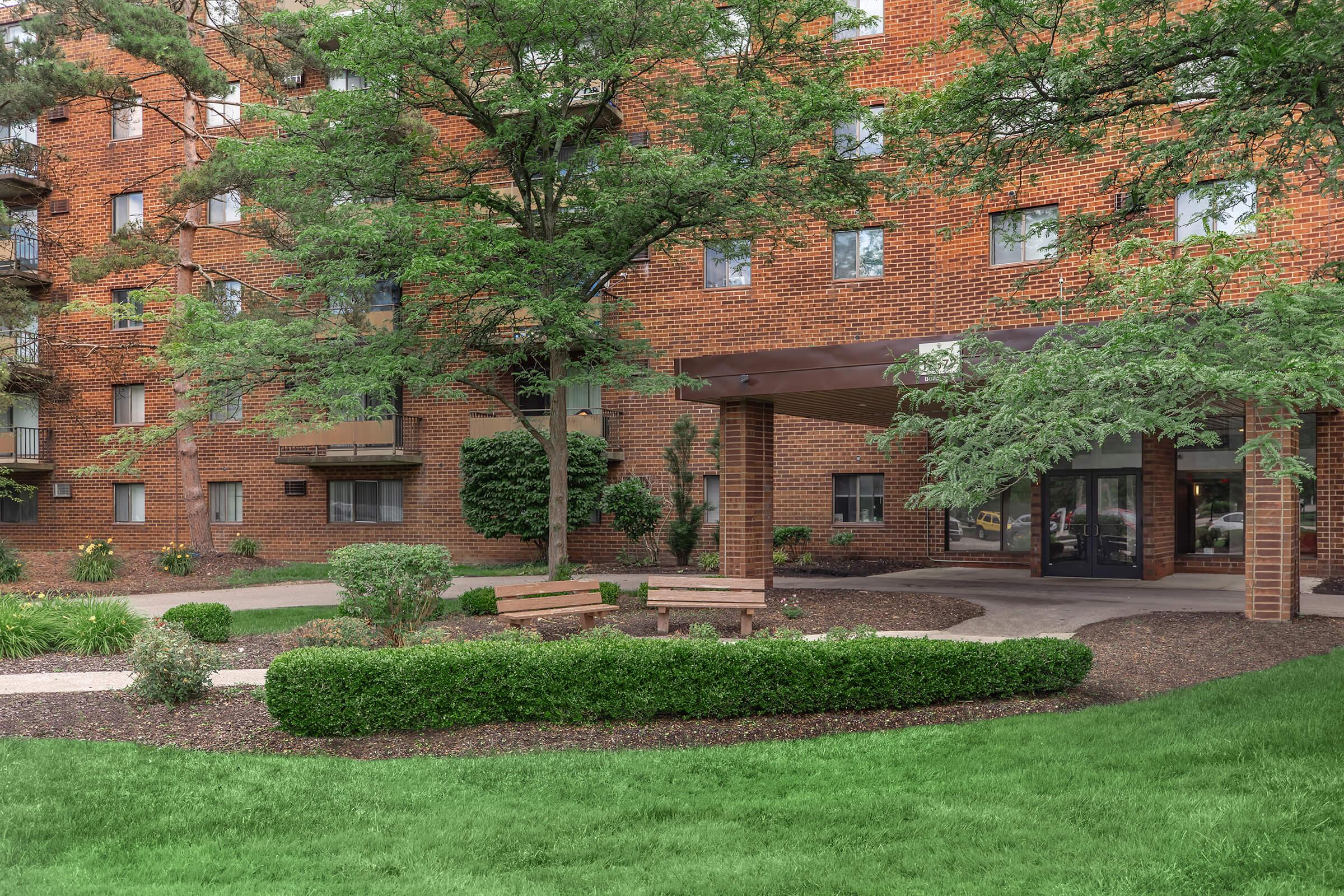 Exterior view of a multi-story brick building surrounded by greenery, including trees and shrubs. A well-maintained lawn features a garden path leading to a covered entrance with benches and flowers, creating a welcoming atmosphere.