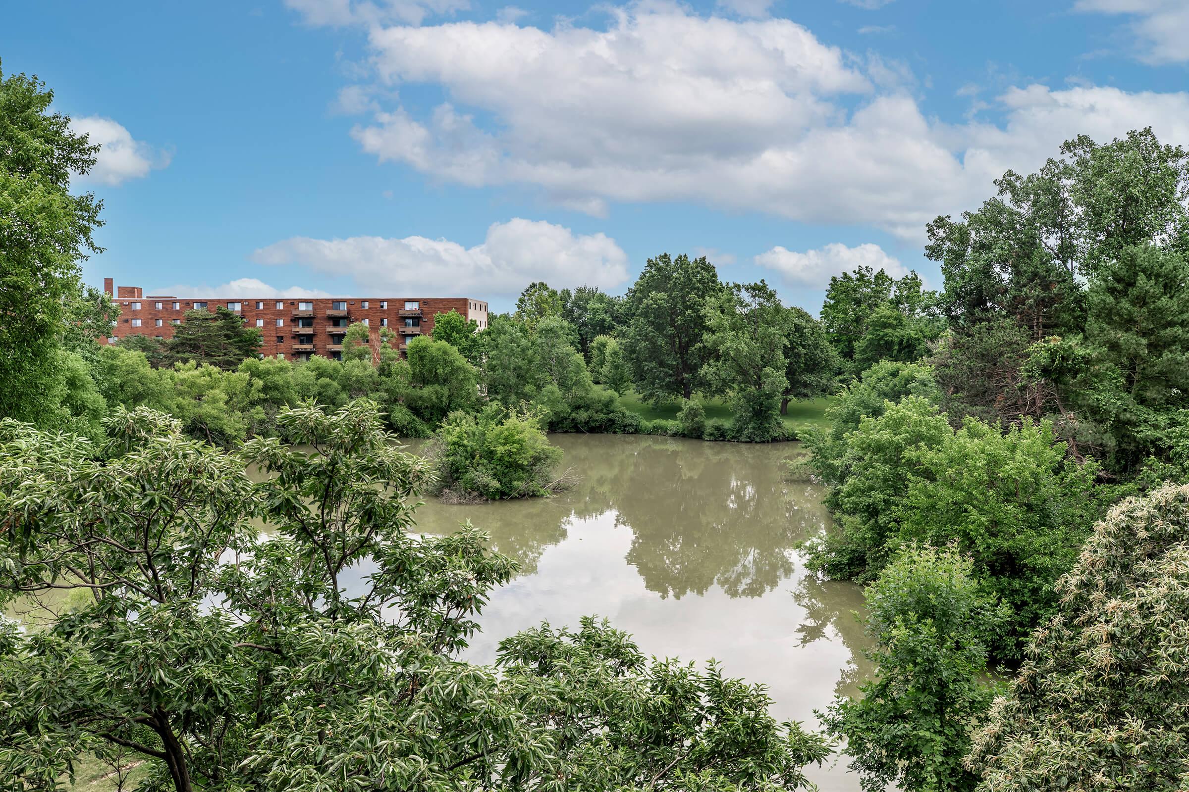A serene view of a calm river surrounded by lush green trees and foliage, reflecting the overcast sky. In the background, a brick apartment building stands amidst the natural landscape, complementing the tranquil setting.