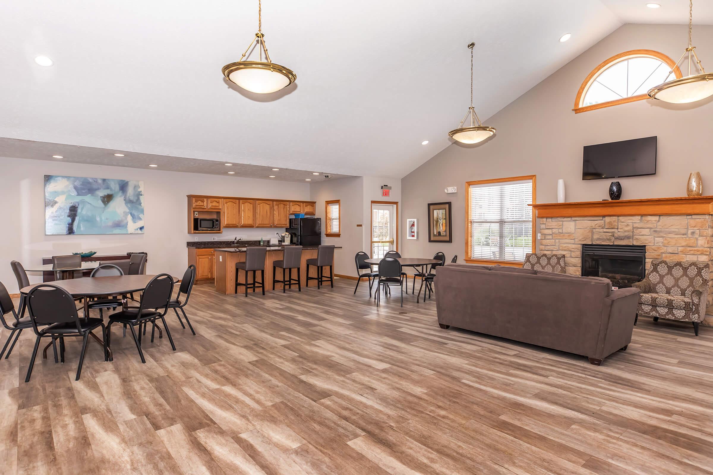 Spacious communal lounge with hardwood-style flooring, featuring a cozy seating area with a sofa, a stone fireplace, and large windows. A kitchen area with wooden cabinets and a dining area with round tables and chairs is visible in the background, creating a welcoming atmosphere for gatherings.
