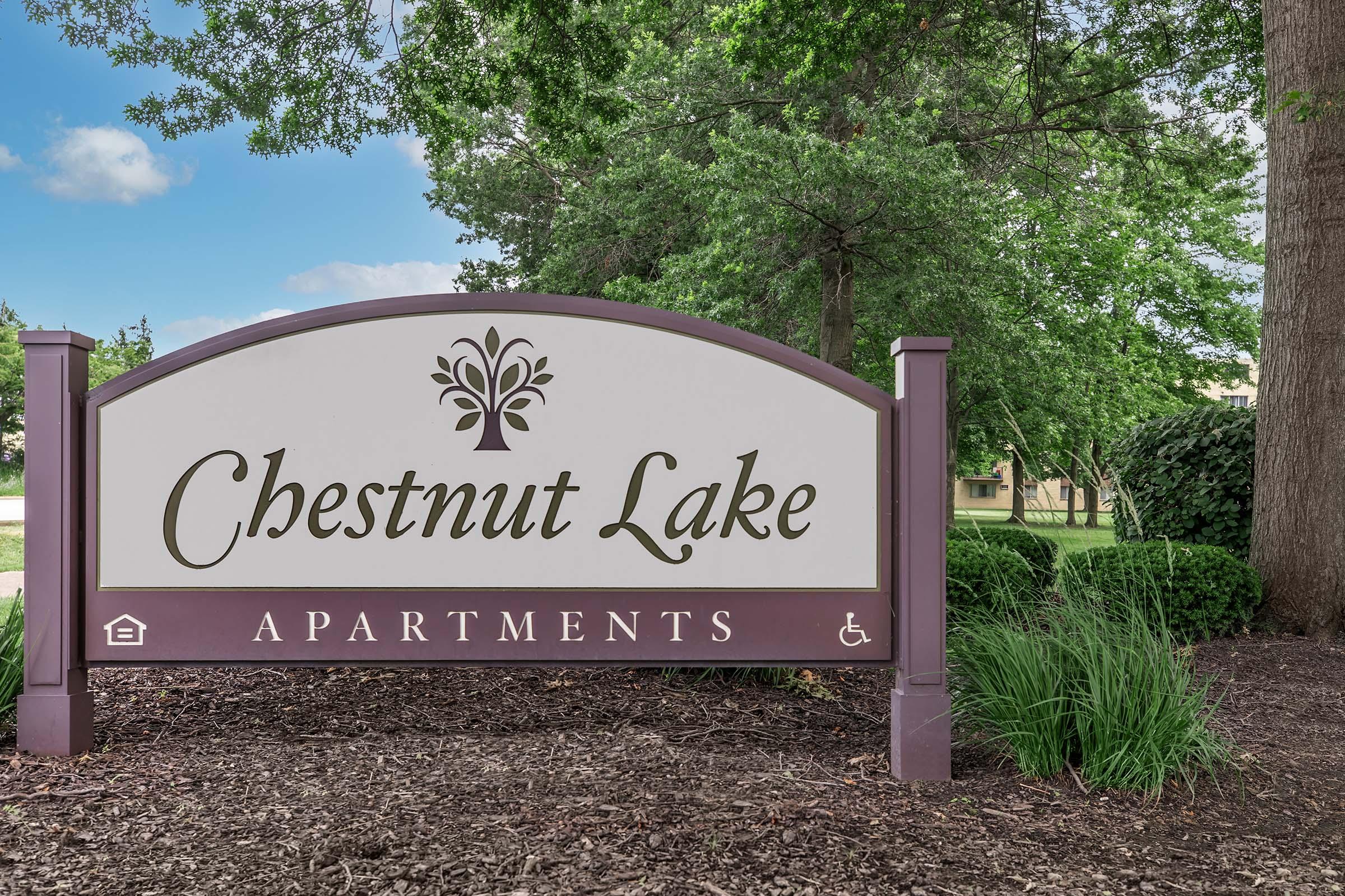 Sign for Chestnut Lake Apartments with a decorative tree logo, surrounded by greenery and trees. The background features a clear blue sky.