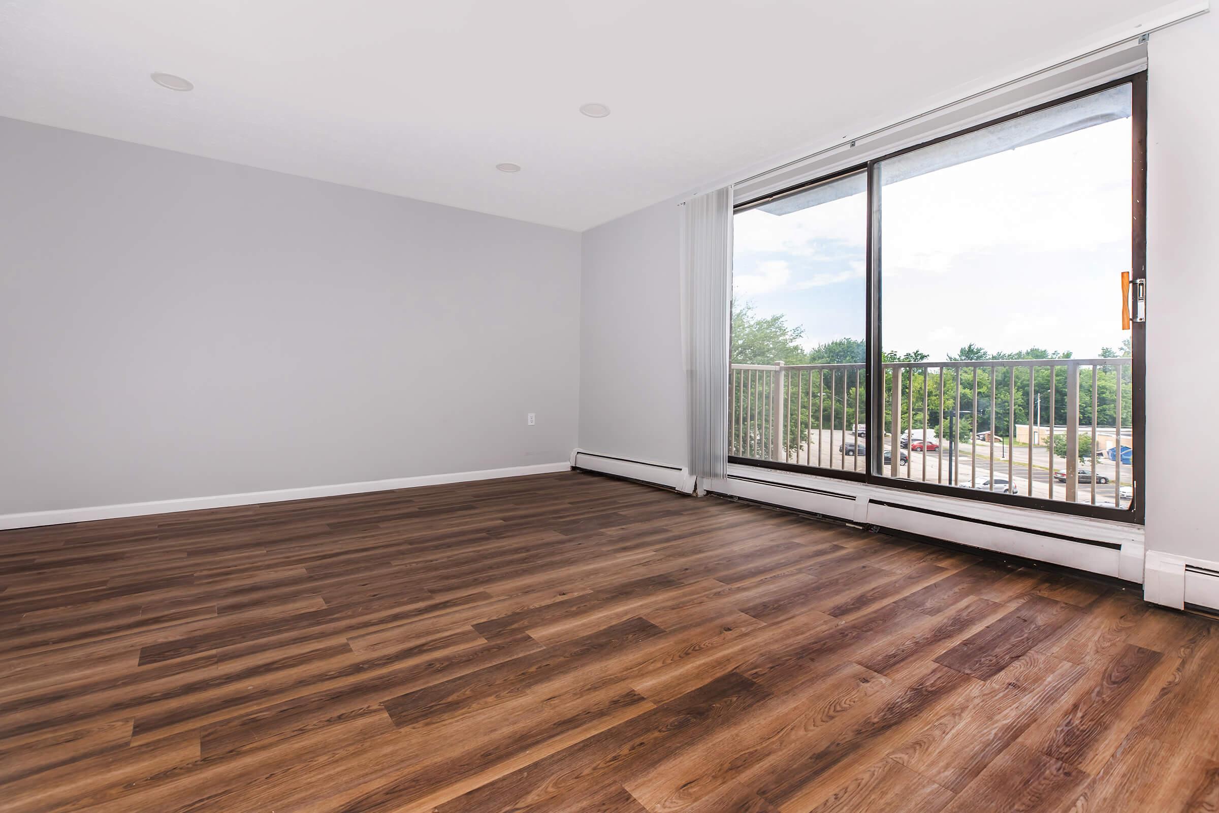 A spacious, unfurnished living room with light gray walls and large windows. The room features hardwood flooring and a view of greenery outside. Natural light brightens the space, and there is a sliding door leading to a small balcony. The overall atmosphere is bright and airy.