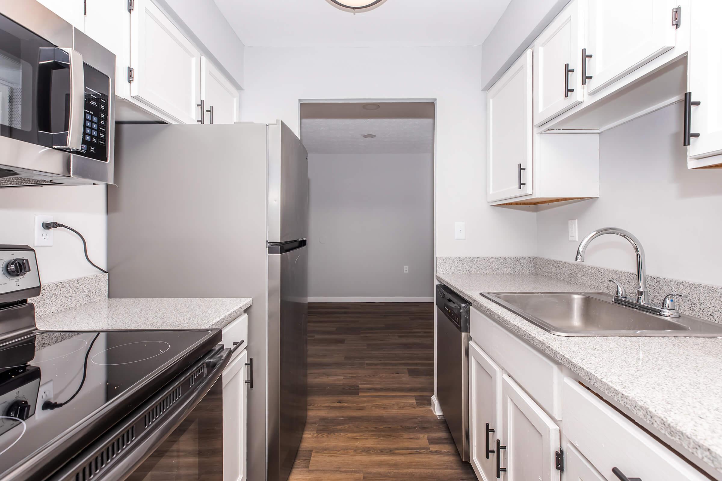 A modern kitchen featuring stainless steel appliances, including a microwave and refrigerator. The countertops are light-colored granite, and there are white cabinets with black handles. A sink is positioned next to the dishwasher, and a doorway leads to another room in the background. The flooring is dark wood.