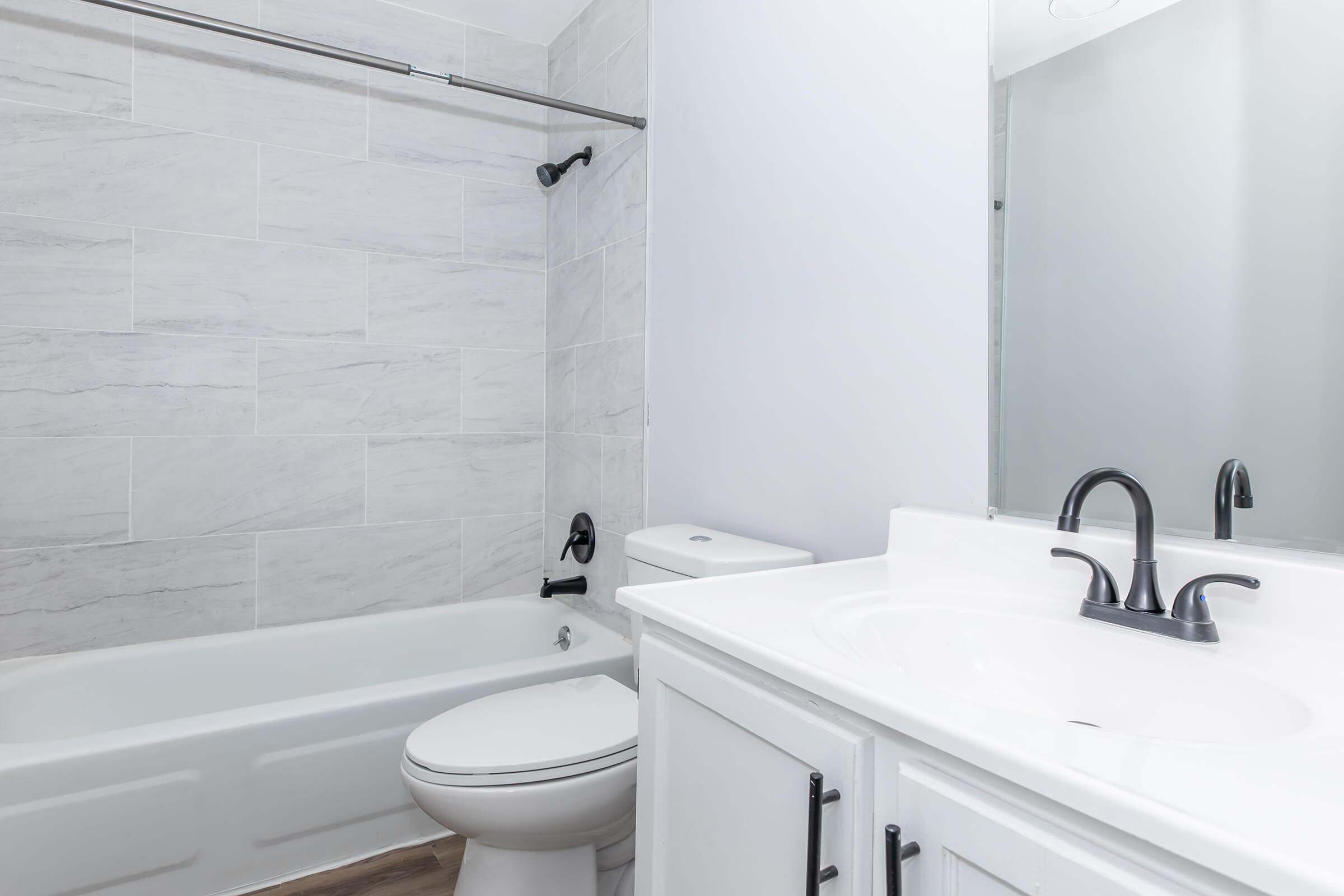 A modern bathroom featuring a white bathtub and toilet, gray tiled walls, and a sleek vanity with a black faucet. The space is well-lit with natural light and has a clean, minimalist design.
