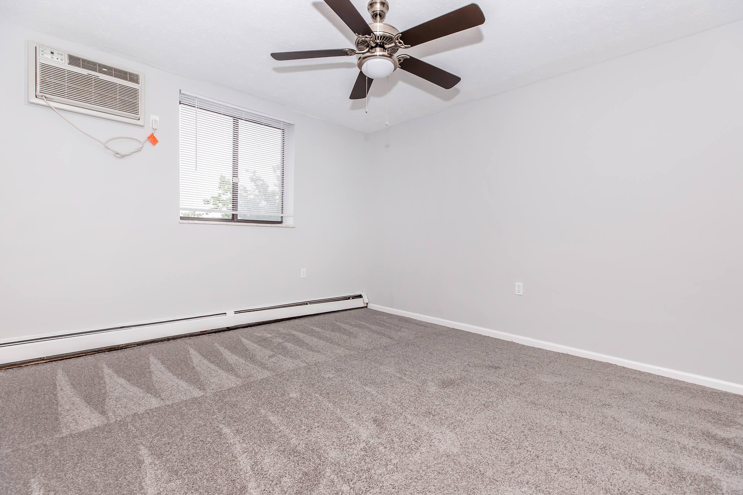 Empty room with light gray walls and a ceiling fan. An air conditioning unit is mounted on the wall near a window with blinds. The floor is covered in soft, light gray carpet, creating a spacious and clean appearance.