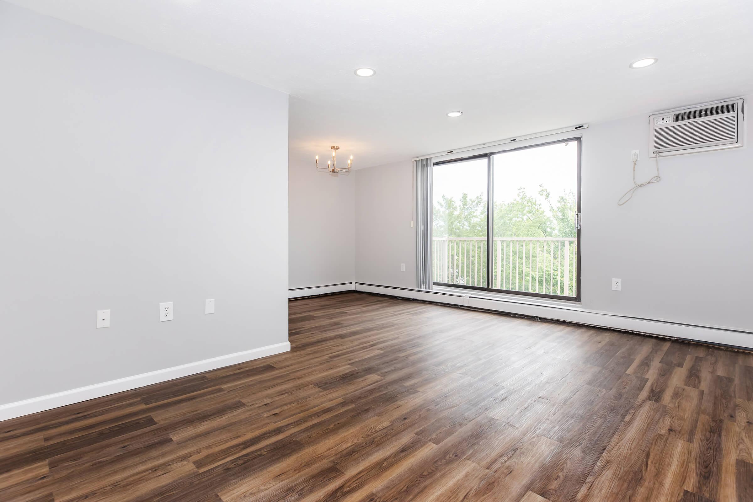 A spacious, modern living room with light gray walls and hardwood flooring. Natural light pours in through a large window with a view of green trees outside. An air conditioning unit is installed near the window, and a simple chandelier hangs from the ceiling, enhancing the contemporary feel of the room.