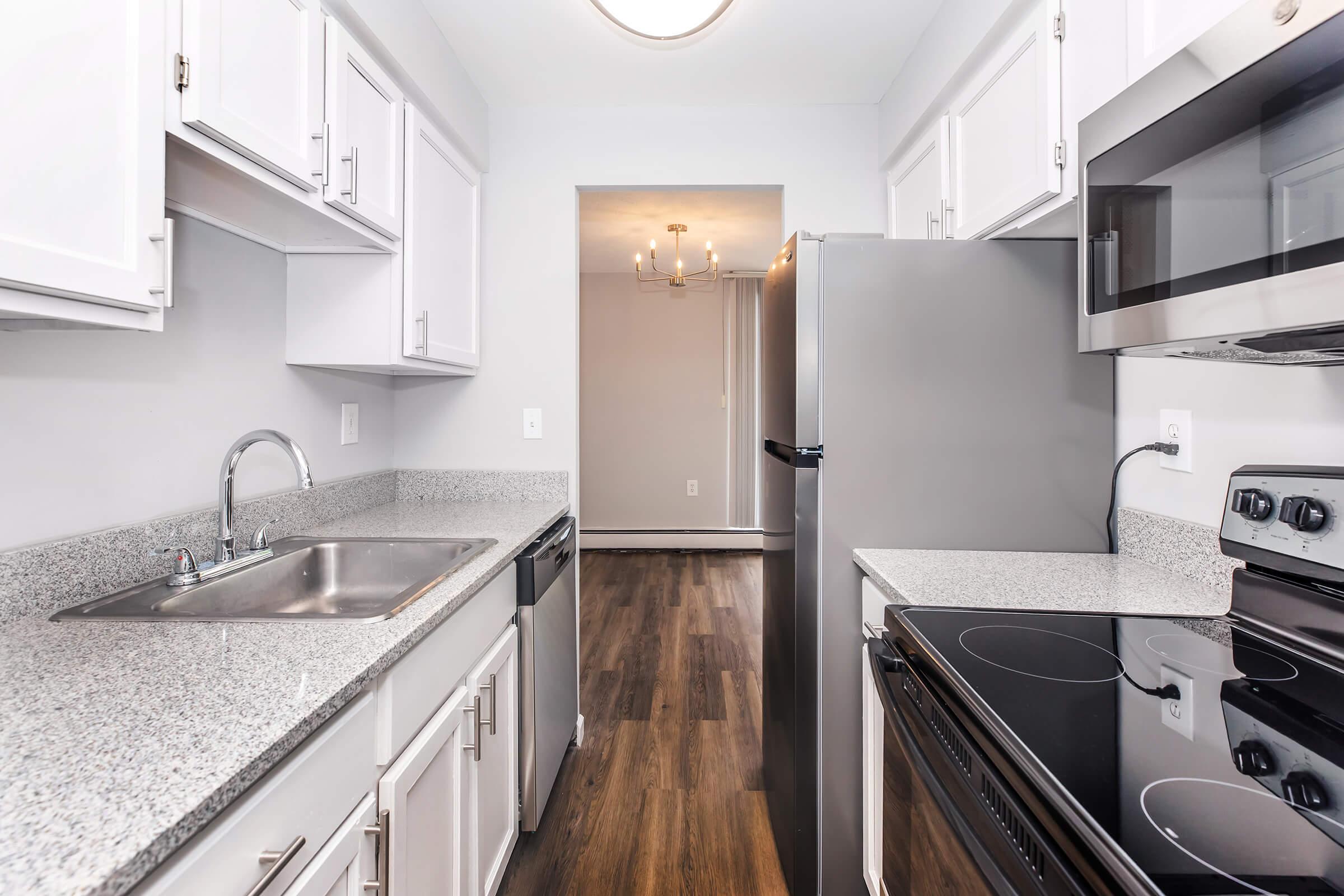 Modern kitchen with white cabinetry, stainless steel appliances including a refrigerator and stove, and a double sink. The countertops are made of gray granite, and the flooring is dark wood. The space is well-lit, featuring a neutral-colored wall and a chandelier in the background.