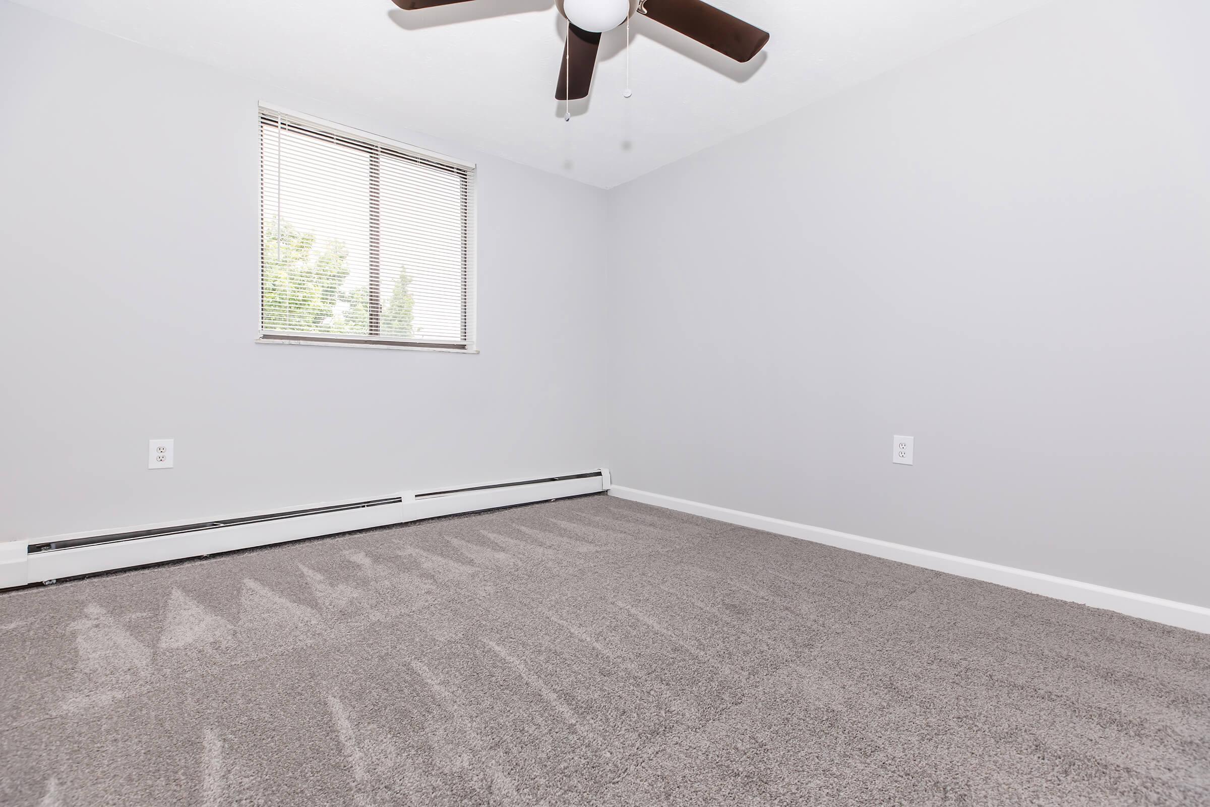 A simple, empty room with light gray walls and a light gray carpet. A ceiling fan is visible, and there's a window with blinds allowing natural light to enter. The room appears clean and spacious, with no furniture present.