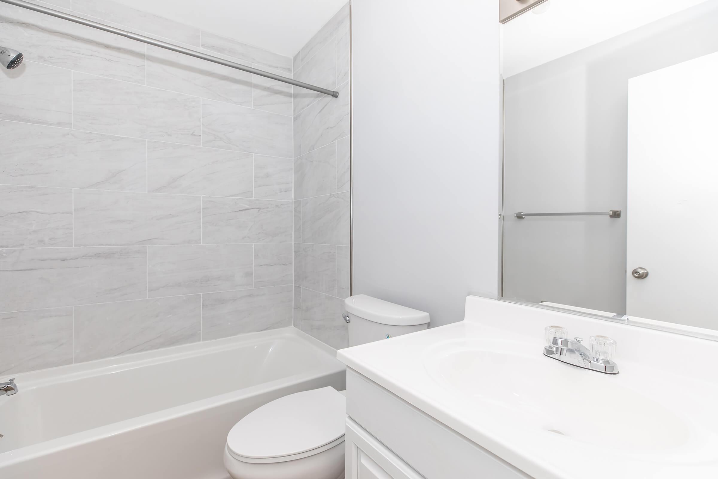 A clean, modern bathroom featuring a bathtub with a shower curtain, a white toilet, and a sink with a mirror above. The walls are tiled in light gray, creating a bright and airy atmosphere. The overall design is minimalist and contemporary.