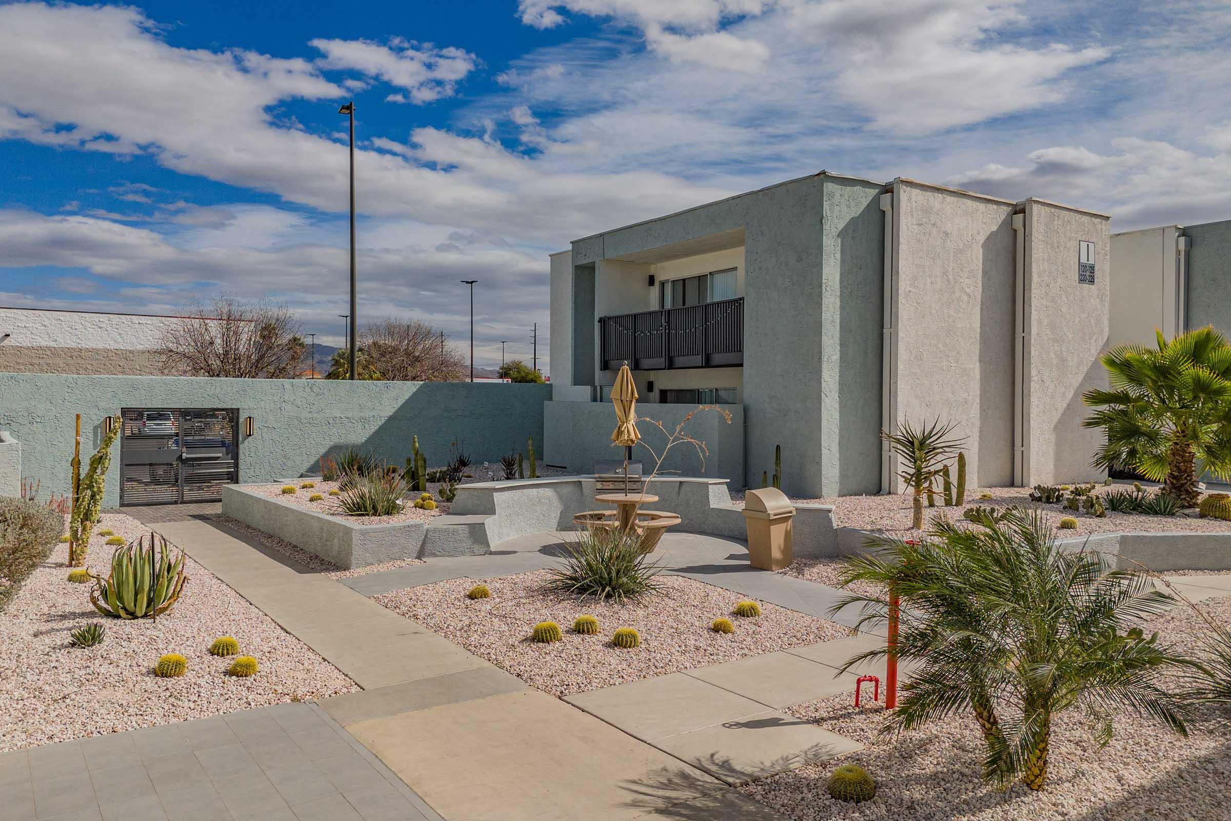 A modern building with a light gray exterior is surrounded by landscaped gardens featuring desert plants, gravel, and a central fountain. Blue skies with scattered clouds provide a backdrop. The pathway leads toward the building entrance, enhancing the serene outdoor atmosphere.