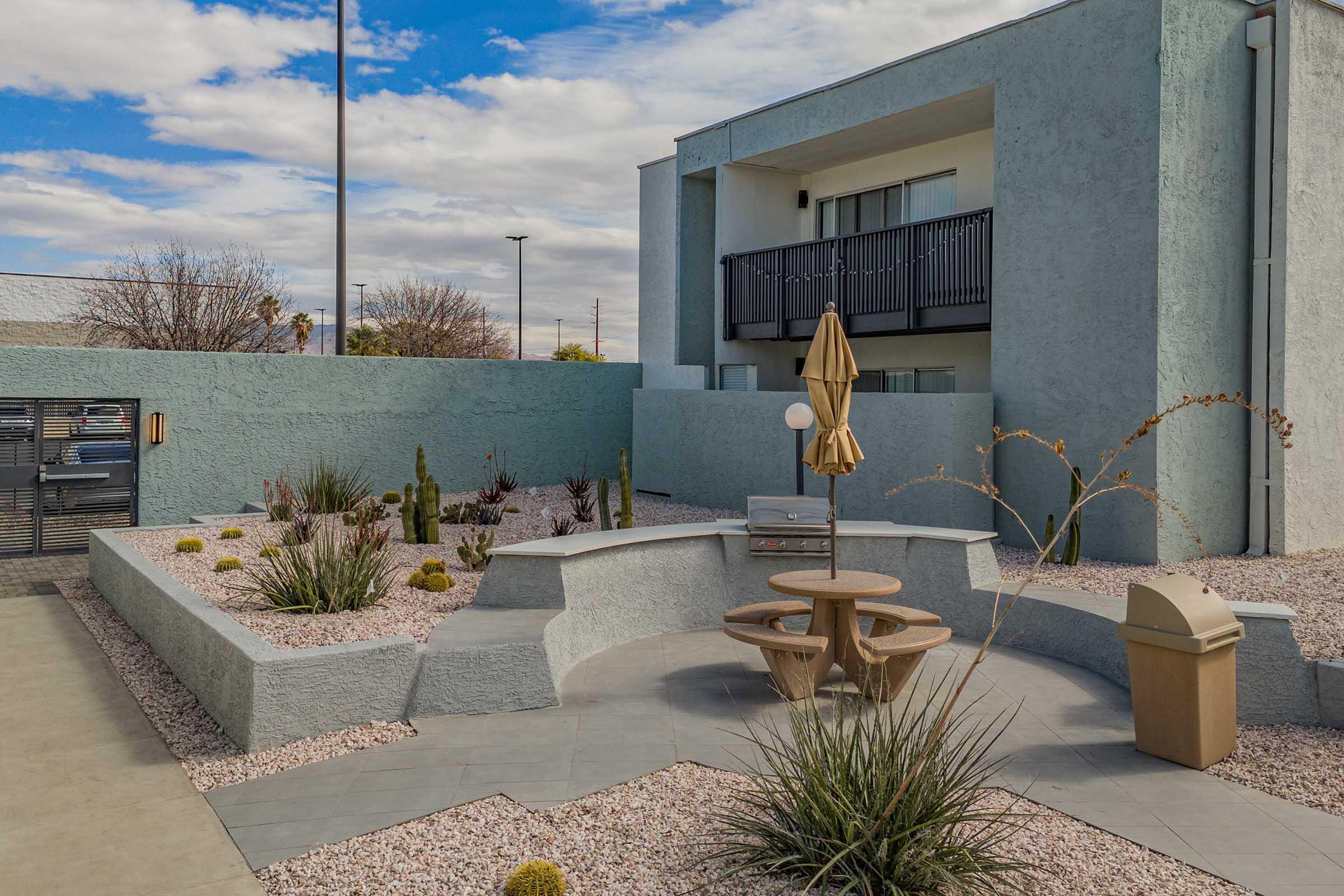 A modern outdoor seating area with a round table and two chairs surrounded by desert landscaping. The space features a gray concrete surface, various plants, an umbrella, and a nearby waste bin. In the background, a building with balconies is visible under a partly cloudy sky.