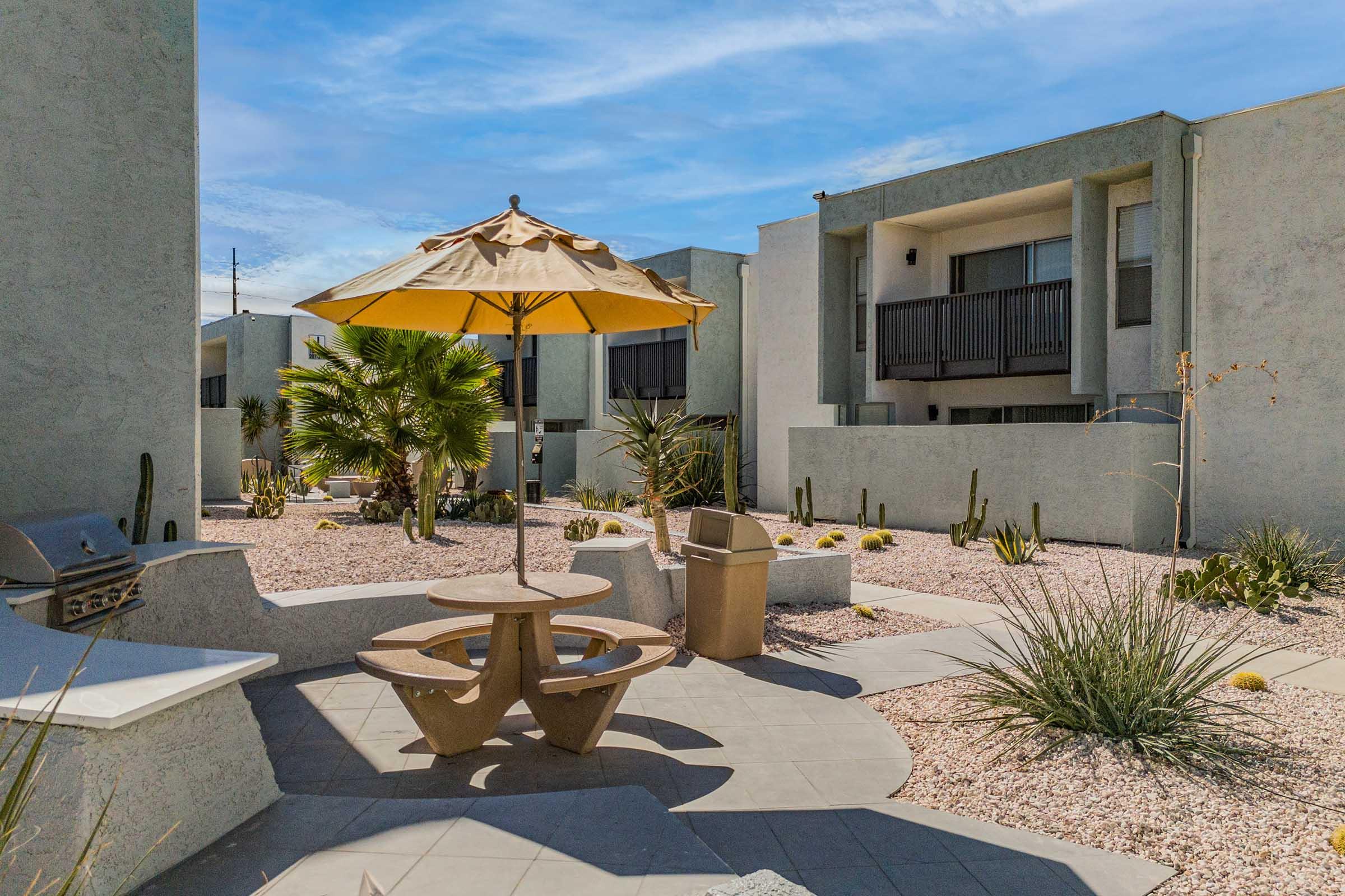 A peaceful courtyard featuring a patio area with a yellow umbrella, a picnic table, and a grill. Surrounding the space are dry landscaping elements, including desert plants and cacti, with nearby apartment buildings in the background under a clear blue sky.