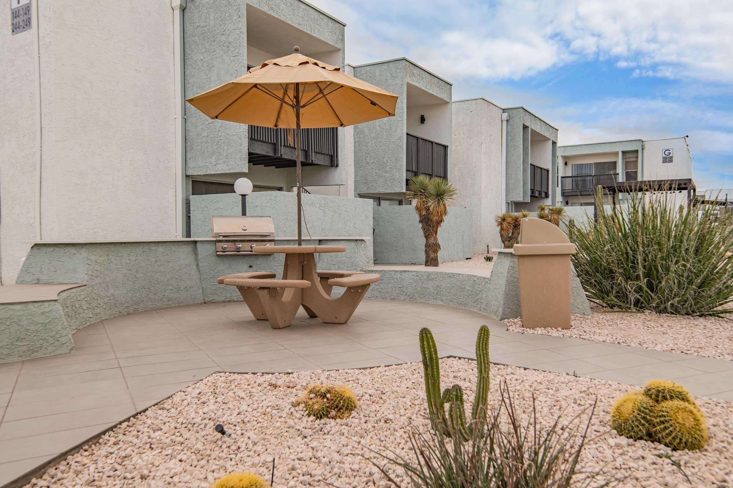 A cozy outdoor seating area featuring a picnic table with an umbrella, surrounded by gravel landscaping and cacti. Behind the seating area, there are modern apartment buildings set against a partly cloudy sky. The scene is tranquil and inviting, perfect for relaxation.