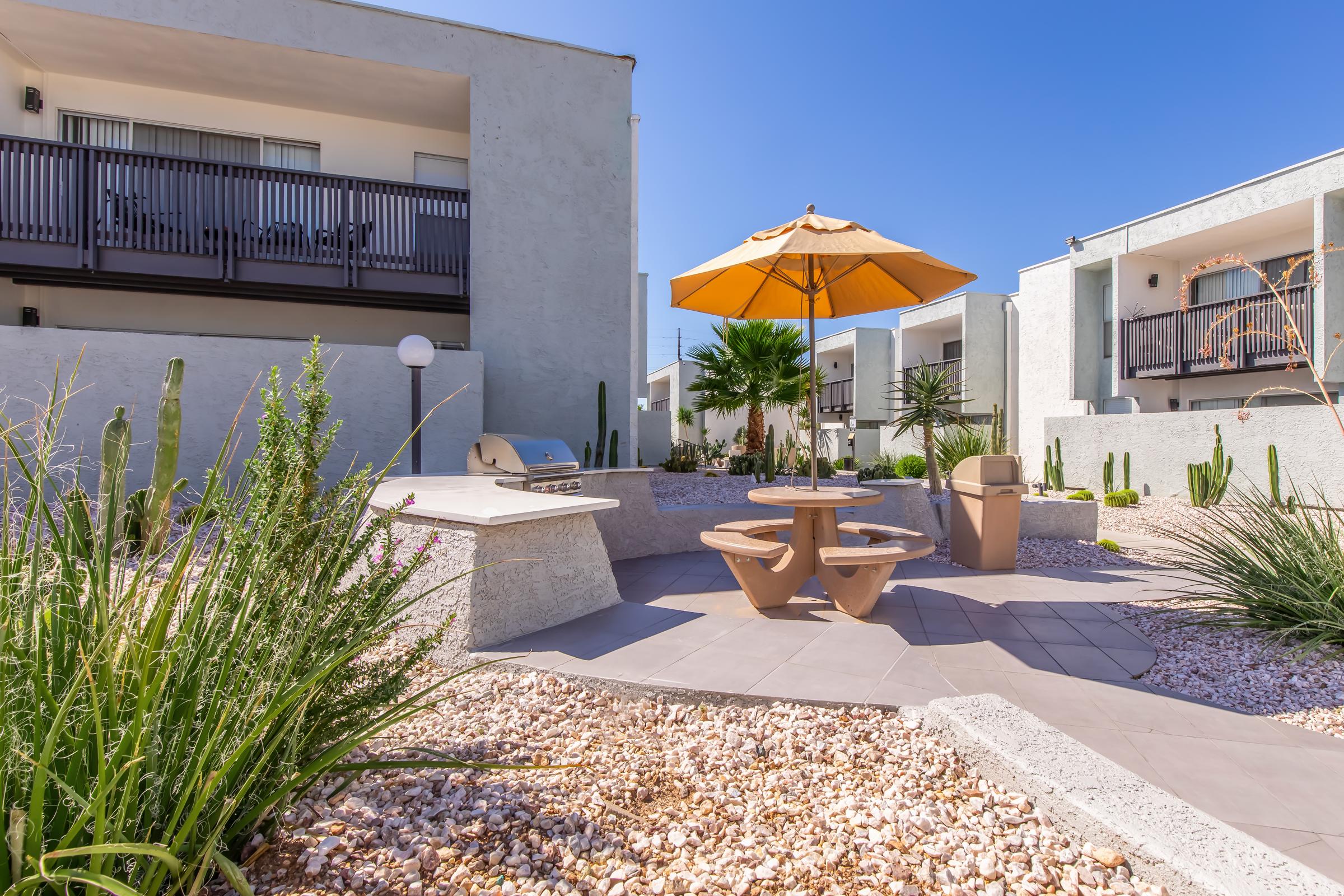 A modern outdoor space featuring a picnic table and umbrella in a landscaped area with cacti and gravel. In the background, there are two multi-story buildings with balconies, and a grill is available for barbecuing. Bright blue sky complements the vibrant setting.