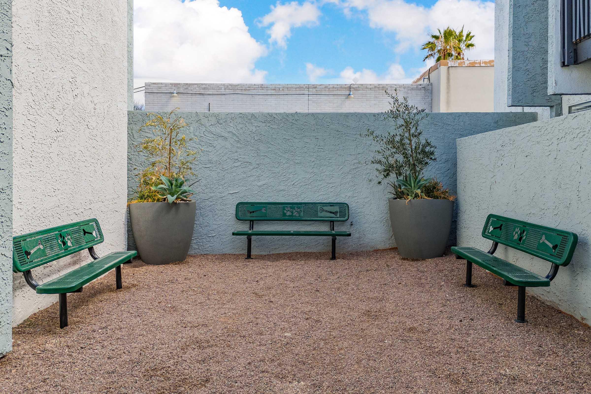 A small outdoor space featuring three green benches arranged around a gravel area, flanked by potted plants. The walls are painted in a light color, and there are scattered clouds in the sky above, creating a serene and inviting atmosphere.