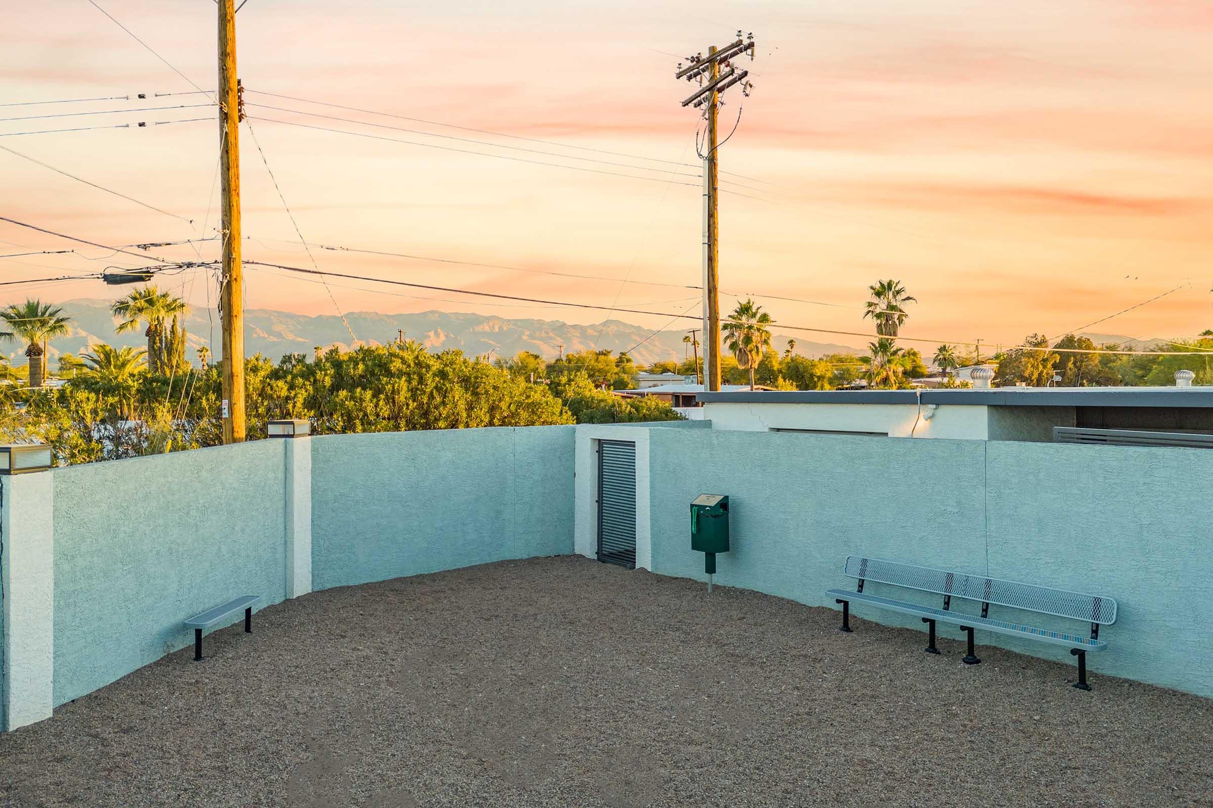 A quiet outdoor space featuring a gravel surface, a green mailbox, and a gray bench. The backdrop includes palm trees and distant mountains under a colorful sunset sky with soft pink and orange hues. Utility poles and power lines frame the scene, adding to the serene ambiance.