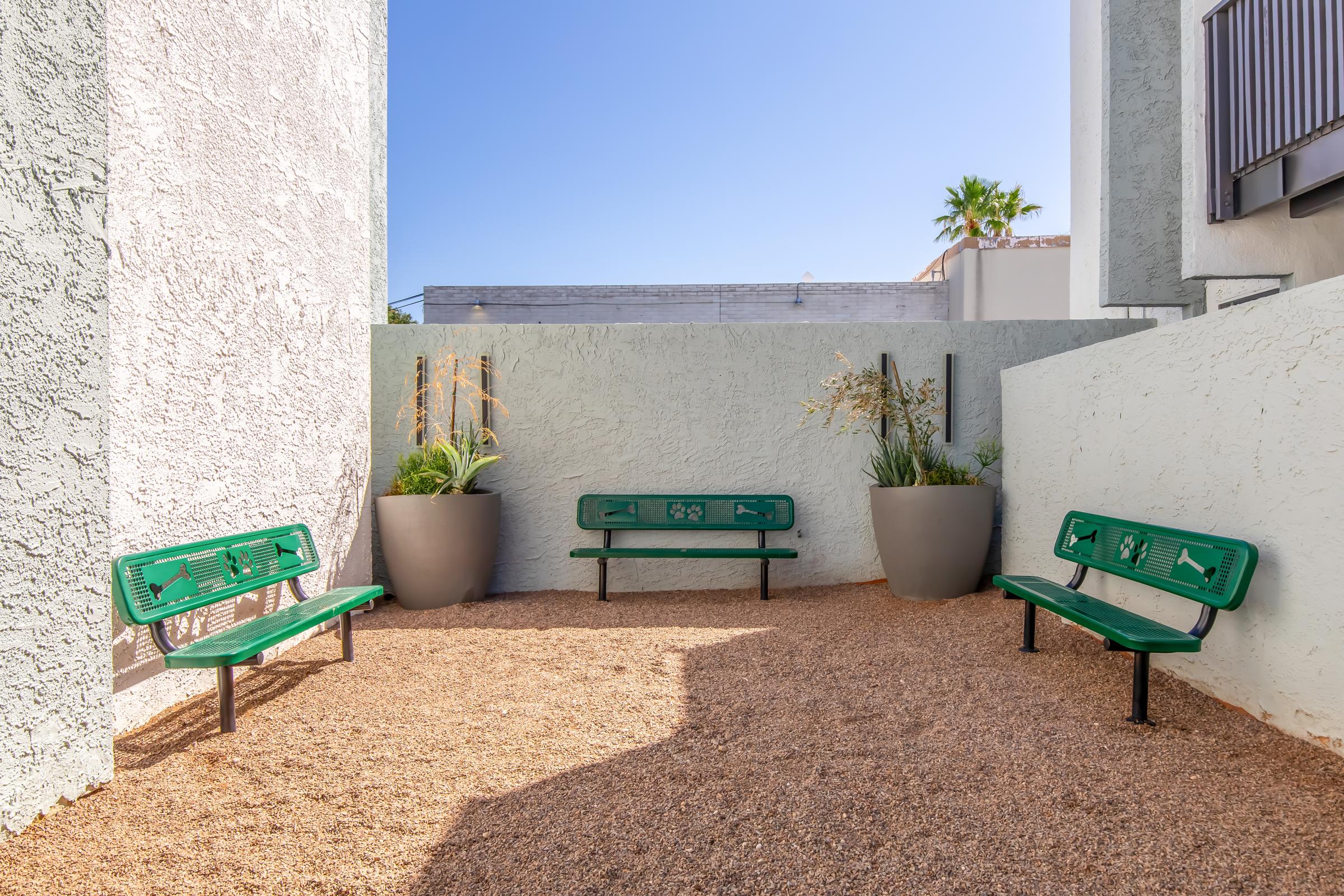 A small outdoor seating area featuring three green metal benches arranged in a circle on a gravel surface. Surrounding the benches are large gray planters with plants. The walls are light-colored, and there is a clear blue sky visible overhead, contributing to a tranquil atmosphere.