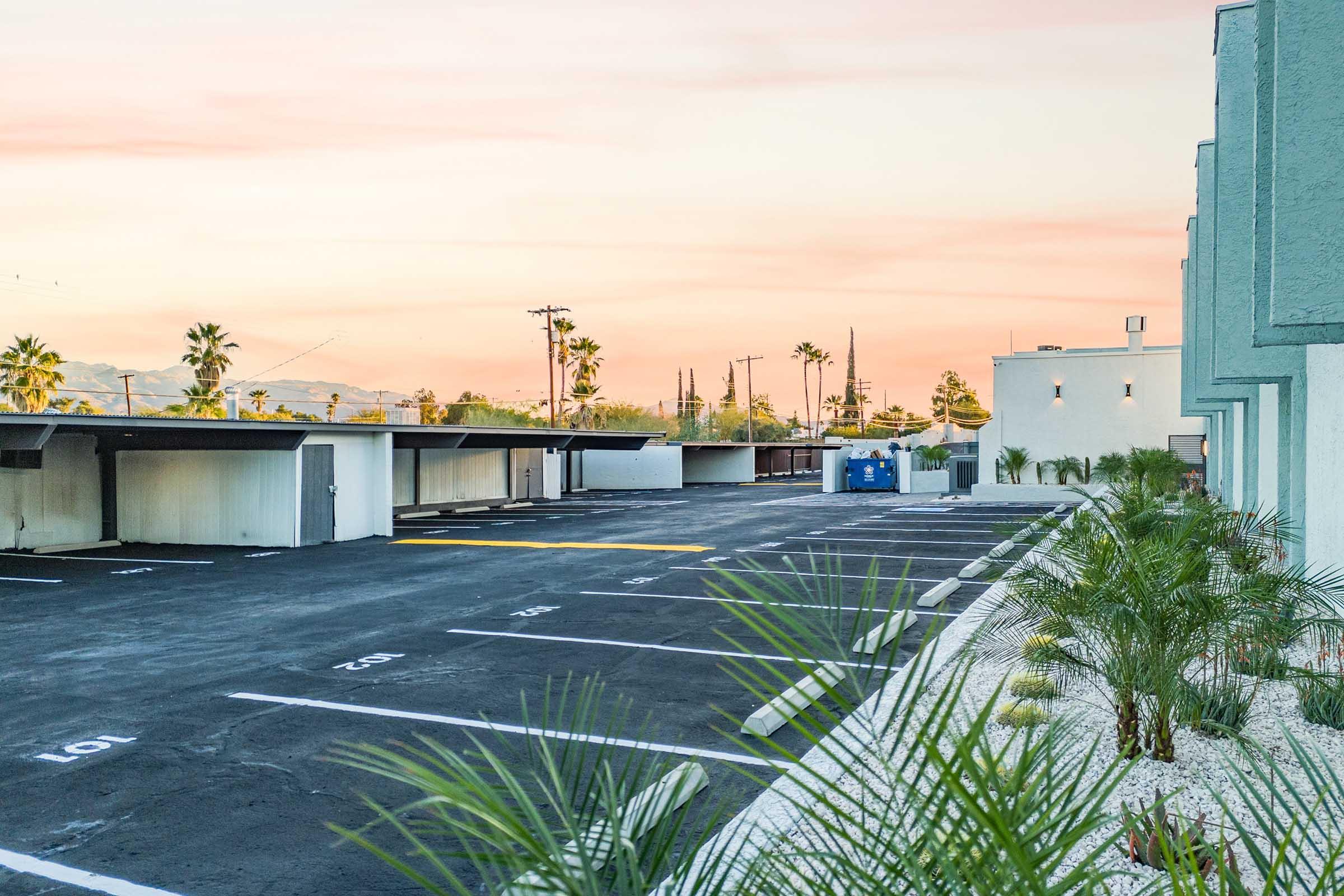 A view of an empty parking lot with marked spaces, surrounded by palm trees and modern buildings under a colorful sunset sky. The scene captures a serene atmosphere with a clear horizon in the background.