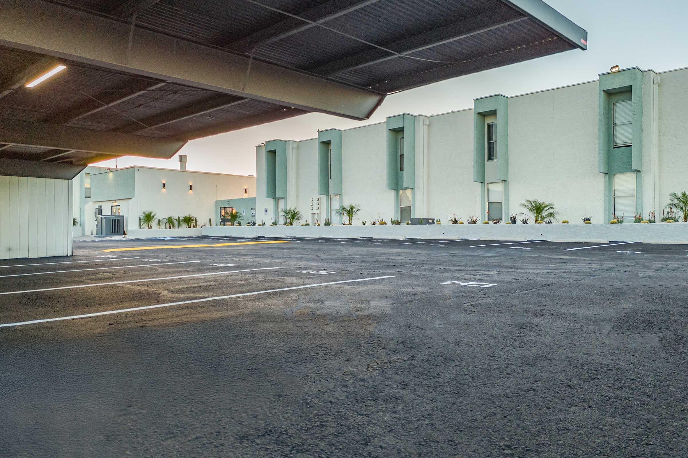 A vacant parking lot beneath a covered structure, with several empty parking spaces. In the background, a low-rise building features a minimalist design with vertical windows and greenery along the facade. The scene is well-lit, suggesting early morning or late afternoon.