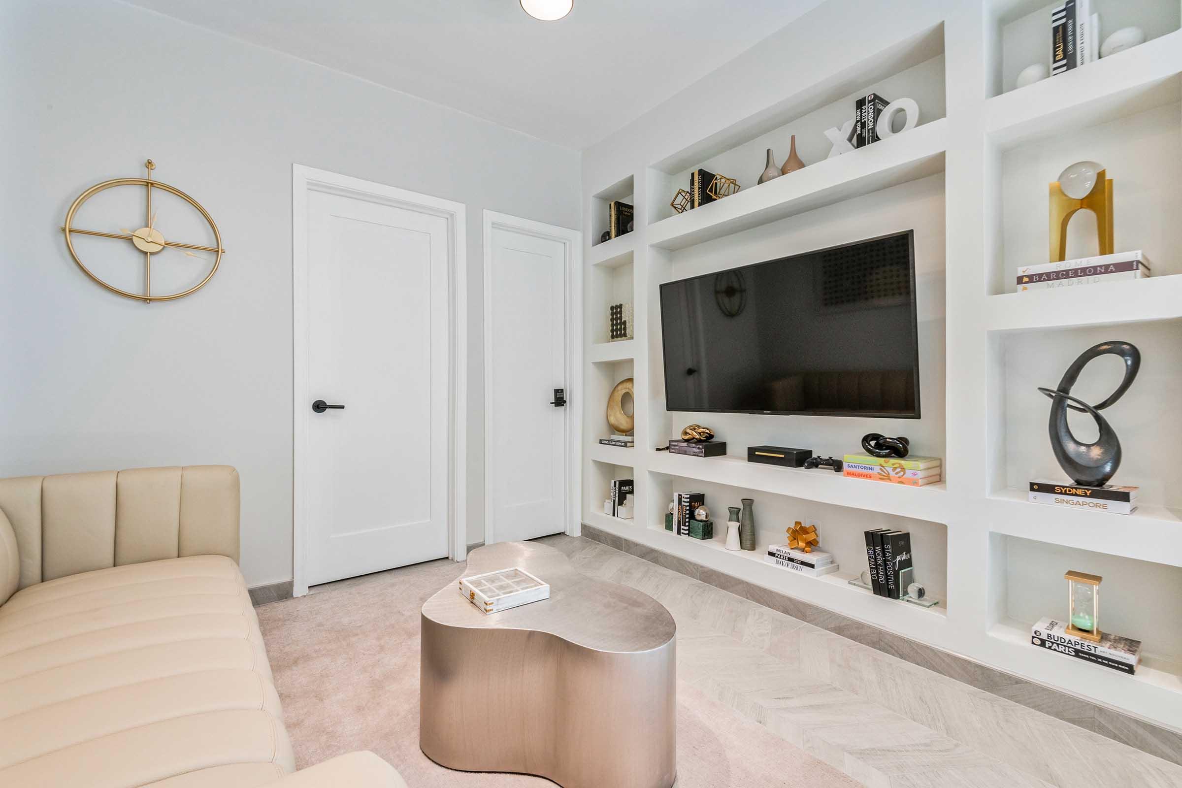 A modern living room featuring a light beige sofa, a sleek, round coffee table, and a built-in shelf with decorative objects and books. A wall-mounted television is placed above the shelf, and there are two closed doors on the right. The floor has a light herringbone pattern, and the walls are painted in a soft gray.