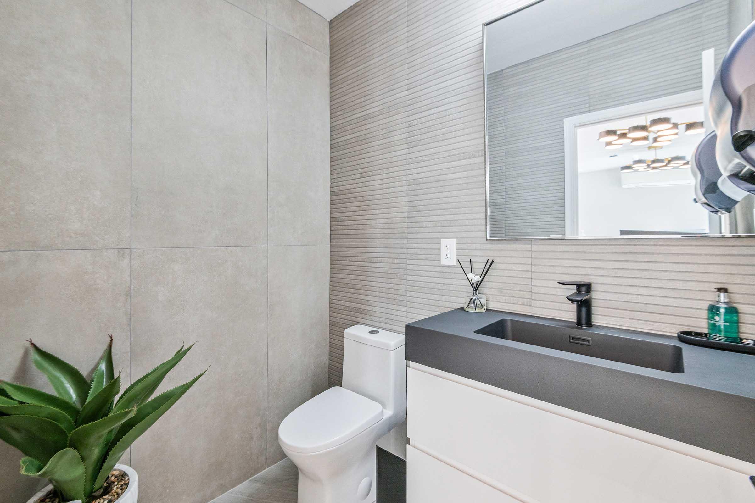 A modern bathroom featuring a sleek gray and beige design. It includes a white toilet, a dark countertop with a sink, a wall-mounted mirror, and an aloe plant. The space is well-lit and has a minimalistic aesthetic, with a hand dryer and soap dispenser visible on the wall.