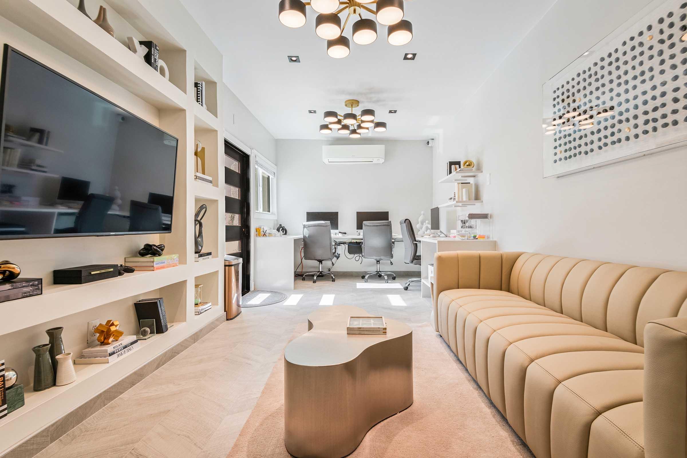 A modern living space featuring a sleek beige sofa, a stylish coffee table, and a wall-mounted television. The decor includes a minimalist bookshelf with decorative items. In the background, there are two office chairs and a desk, creating a blend of relaxation and workspace in a bright, contemporary environment.