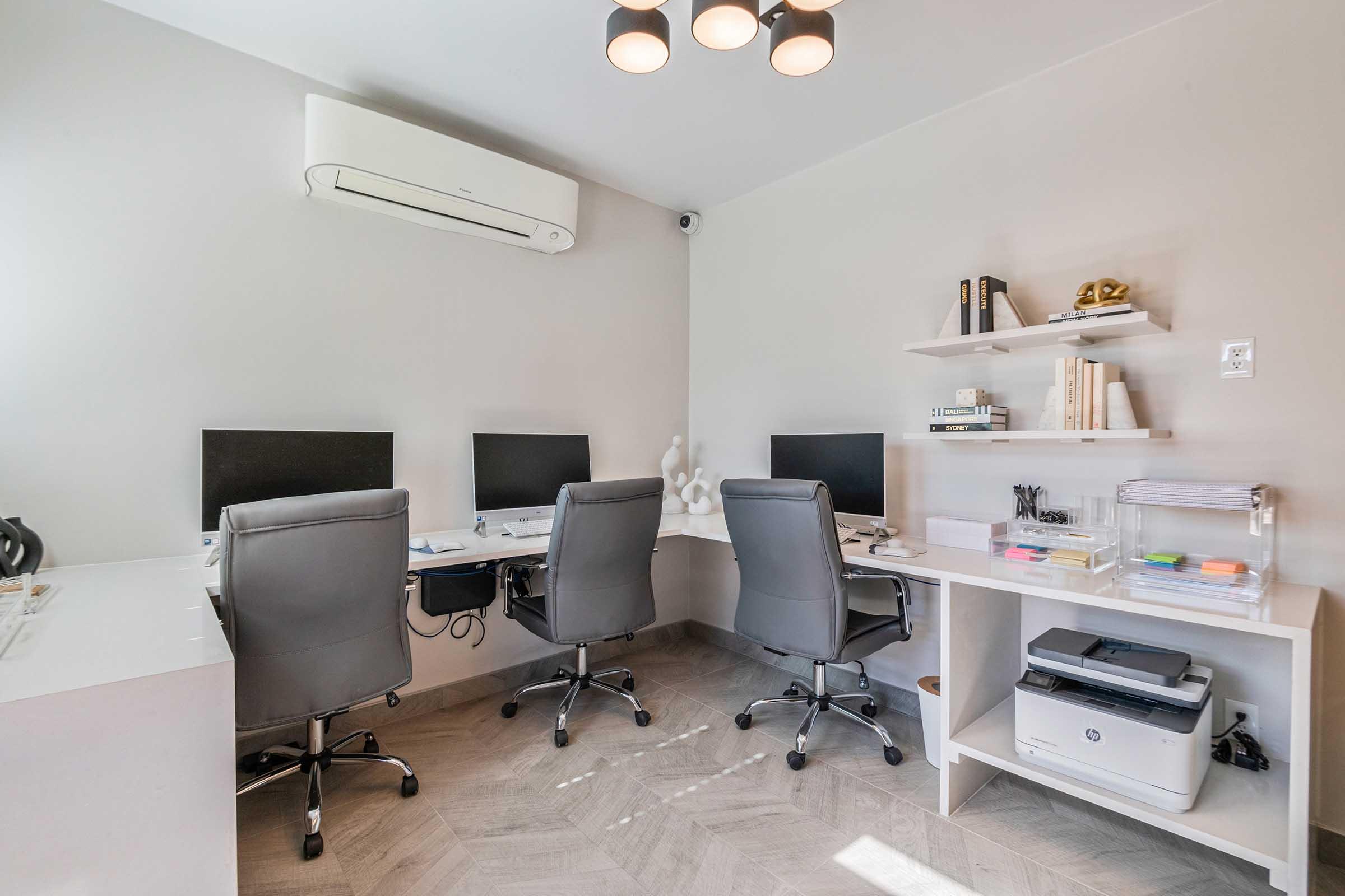 A modern office space featuring two desks with computers, ergonomic chairs, and a shelf with books and decorative items. The walls are painted in a light color, and the room has a sleek, minimalist design. An air conditioning unit is mounted on the wall, providing a clean and organized work environment.