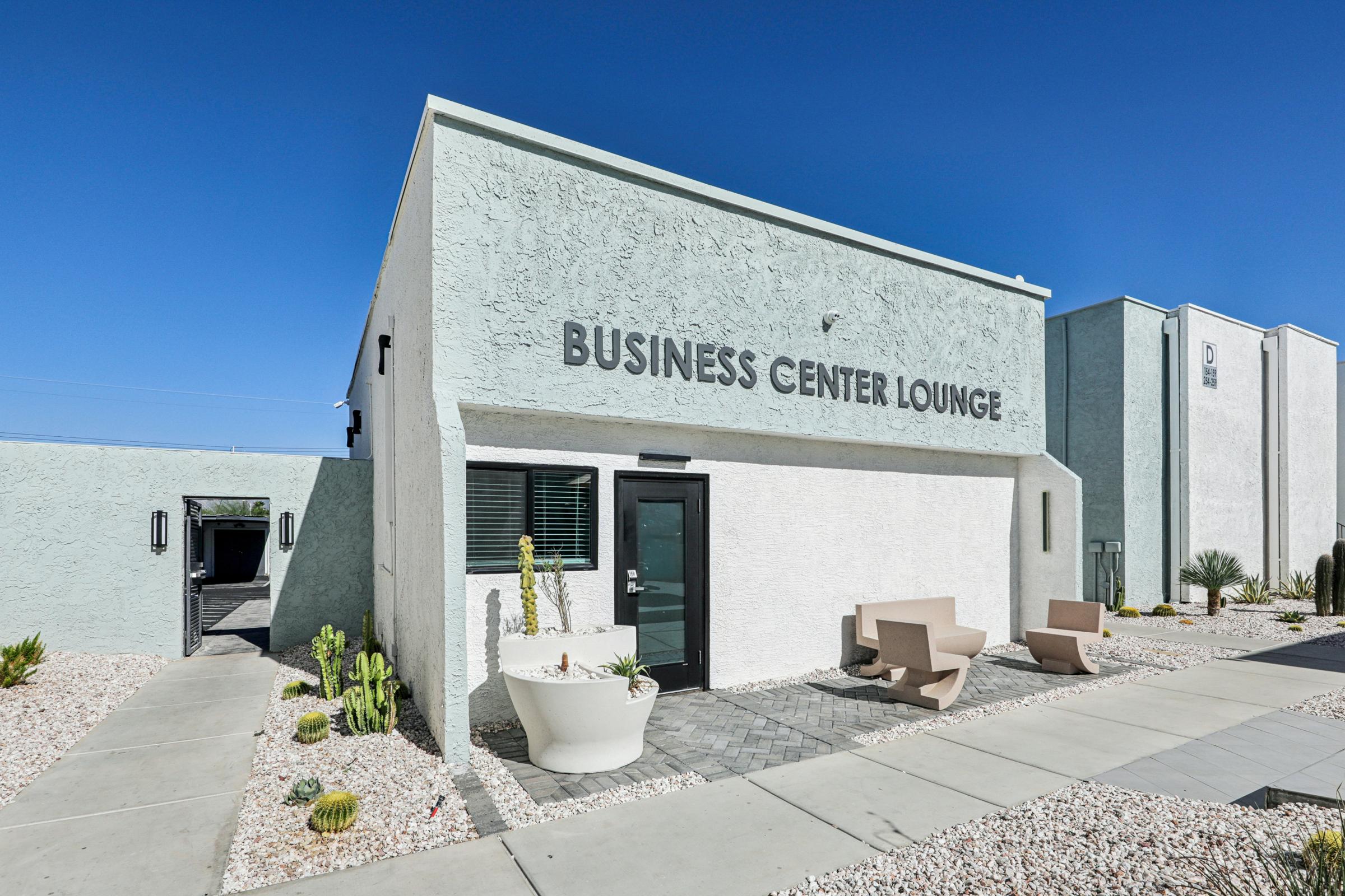 A modern business center lounge with a light-colored exterior. The building features large windows, a black entrance door, and a sign that reads "BUSINESS CENTER LOUNGE." The landscaping includes gravel and potted plants, with a clear blue sky in the background.