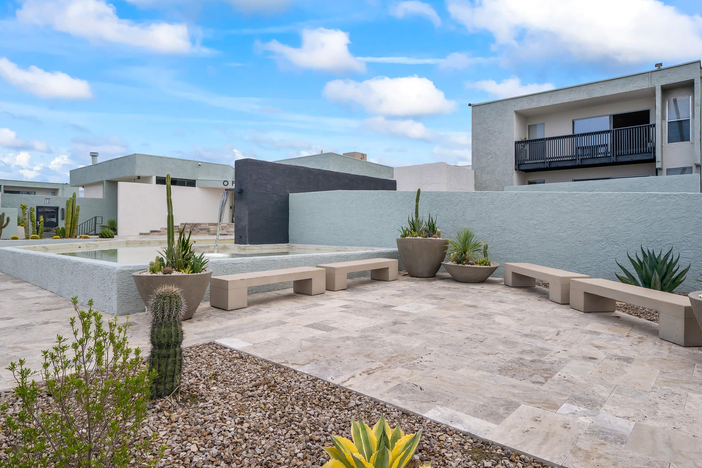 A modern outdoor pool area featuring a shallow water feature surrounded by minimalist landscaping. Large planters with various plants are placed on a stone patio, which includes unique bench seating. The backdrop consists of contemporary buildings under a cloudy blue sky.