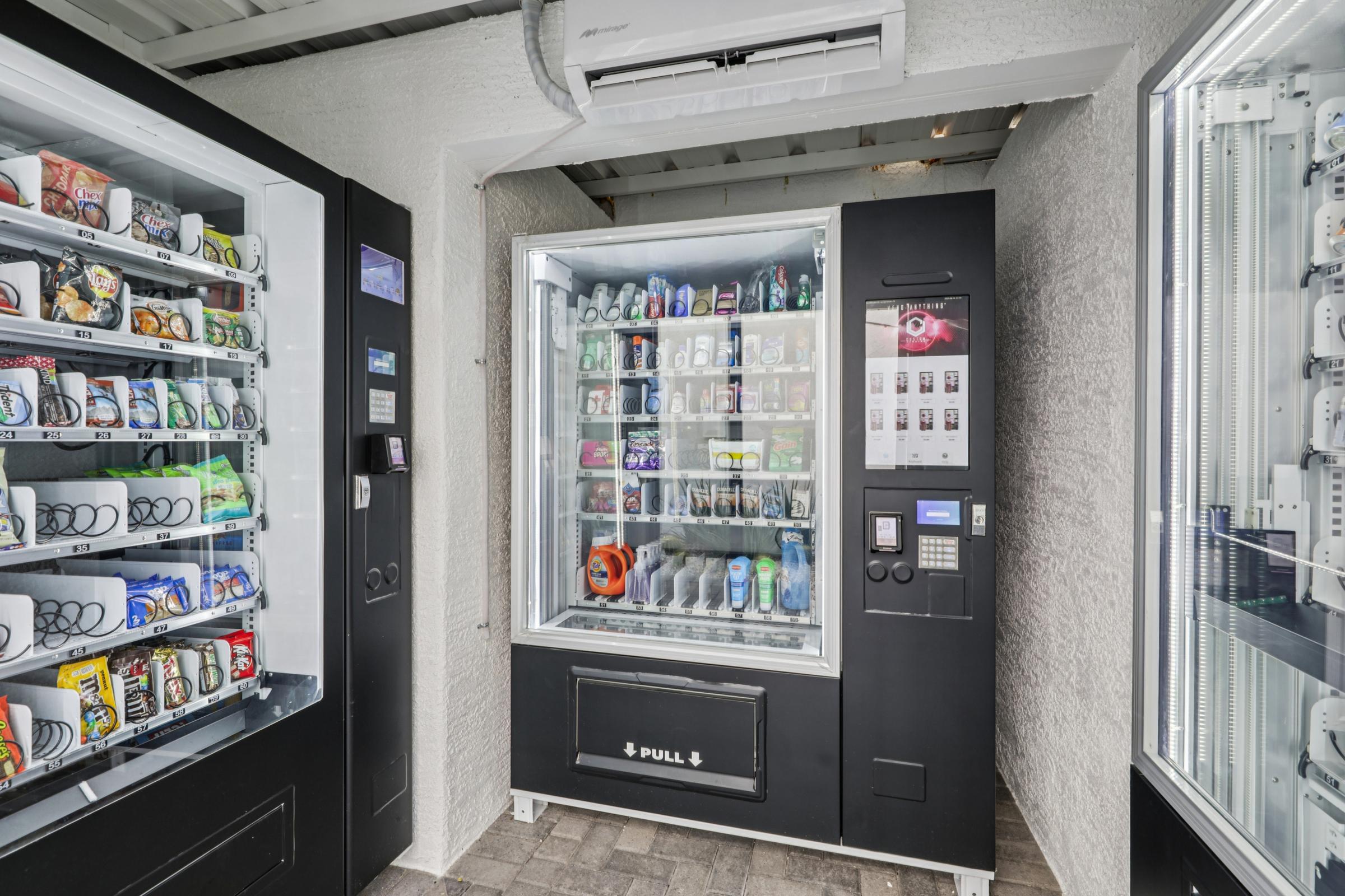A vending machine area featuring two vending machines. The left machine is stocked with various snacks and drinks, while the right machine displays a variety of beverages. The environment is bright and modern, with a white wall and tiled floor. There are buttons and a display panel for purchasing items.