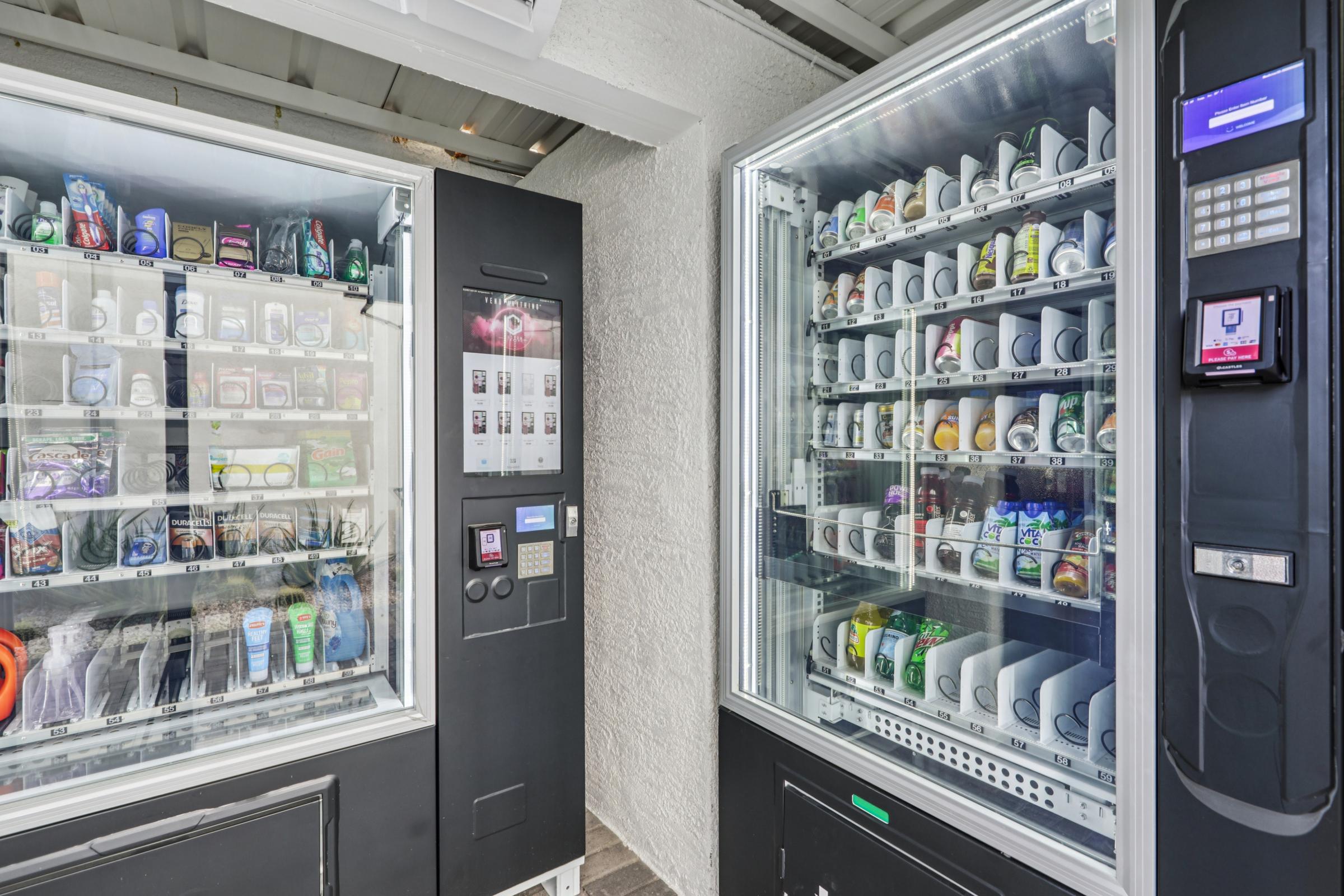 Two vending machines side by side, one containing snacks and drinks while the other offers beverages. The machines have transparent doors showcasing their contents, and a digital keypad for payments is visible on both. The setting appears to be indoors with neutral-colored walls.